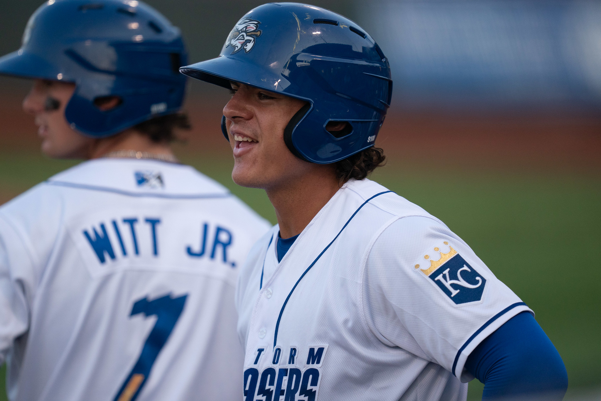 A smiling baseball player (Nick Pratto) is in the foreground in a white jersey top and a blue batting helmet. Teammate Bobby Witt, Jr. is in the background with his back turned and his last name visible on his jersey.