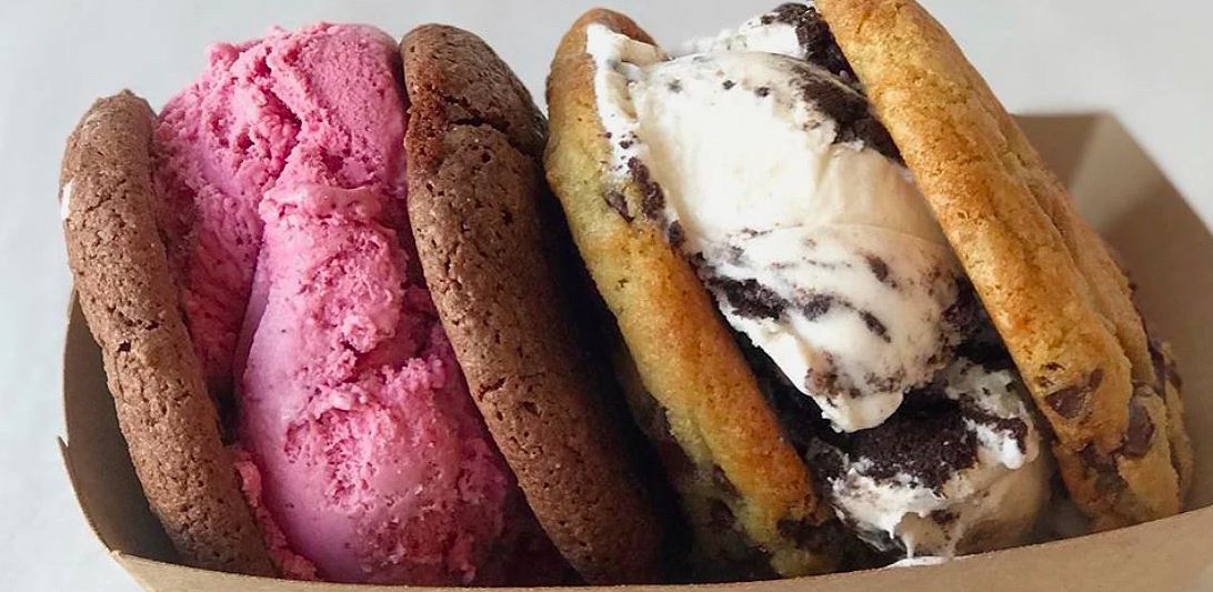 Two cookie ice cream sandwiches sit side by side in a white paper container. One has a pinkish red ice cream in it, while the other has a white ice cream with crumbled Oreos in it.