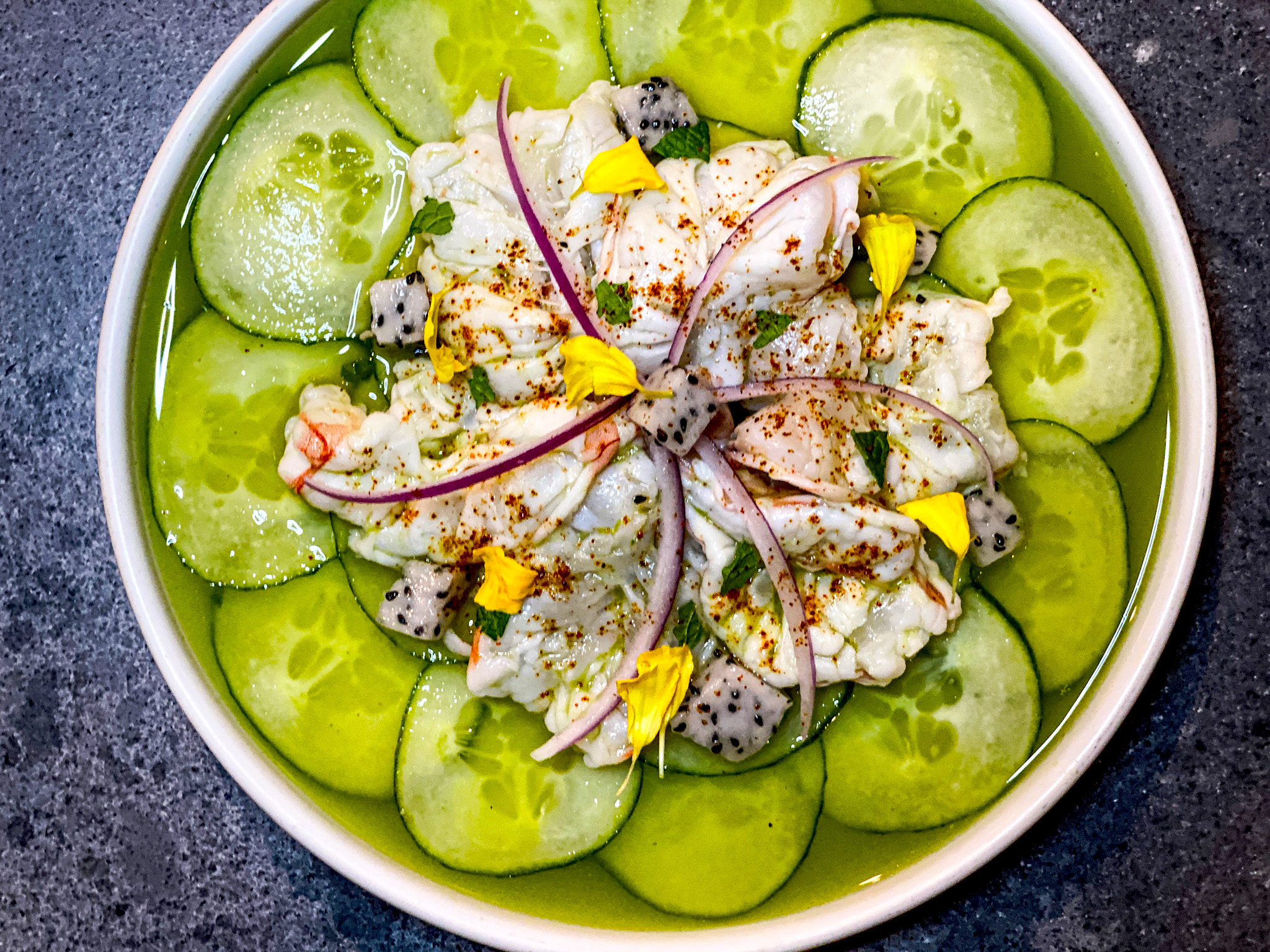 A pile of shrimp ceviche garnished with colorful flecks of vegetables sits atop a concentric circle of cucumber slices 