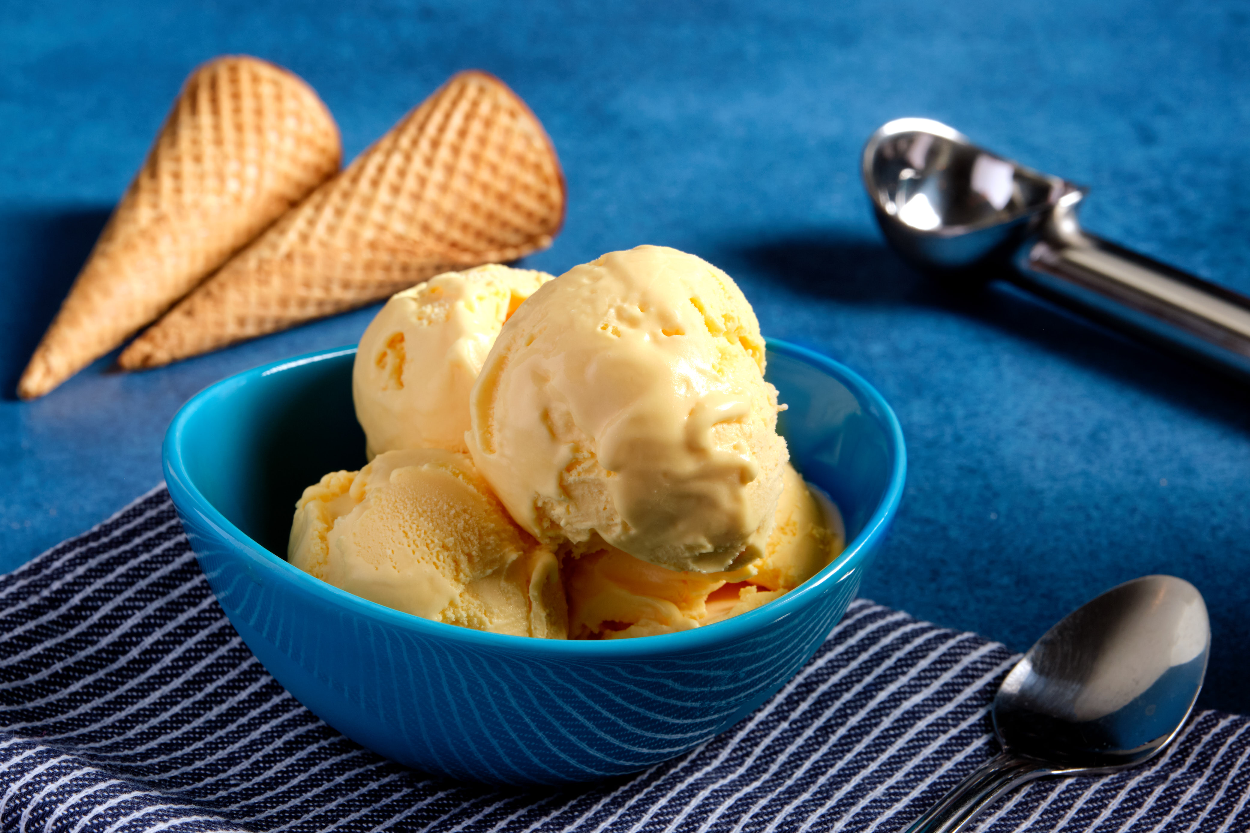 Four scoops of of mac and cheese flavored ice cream in a blue bowl, next to an ice cream scoop and two cones.