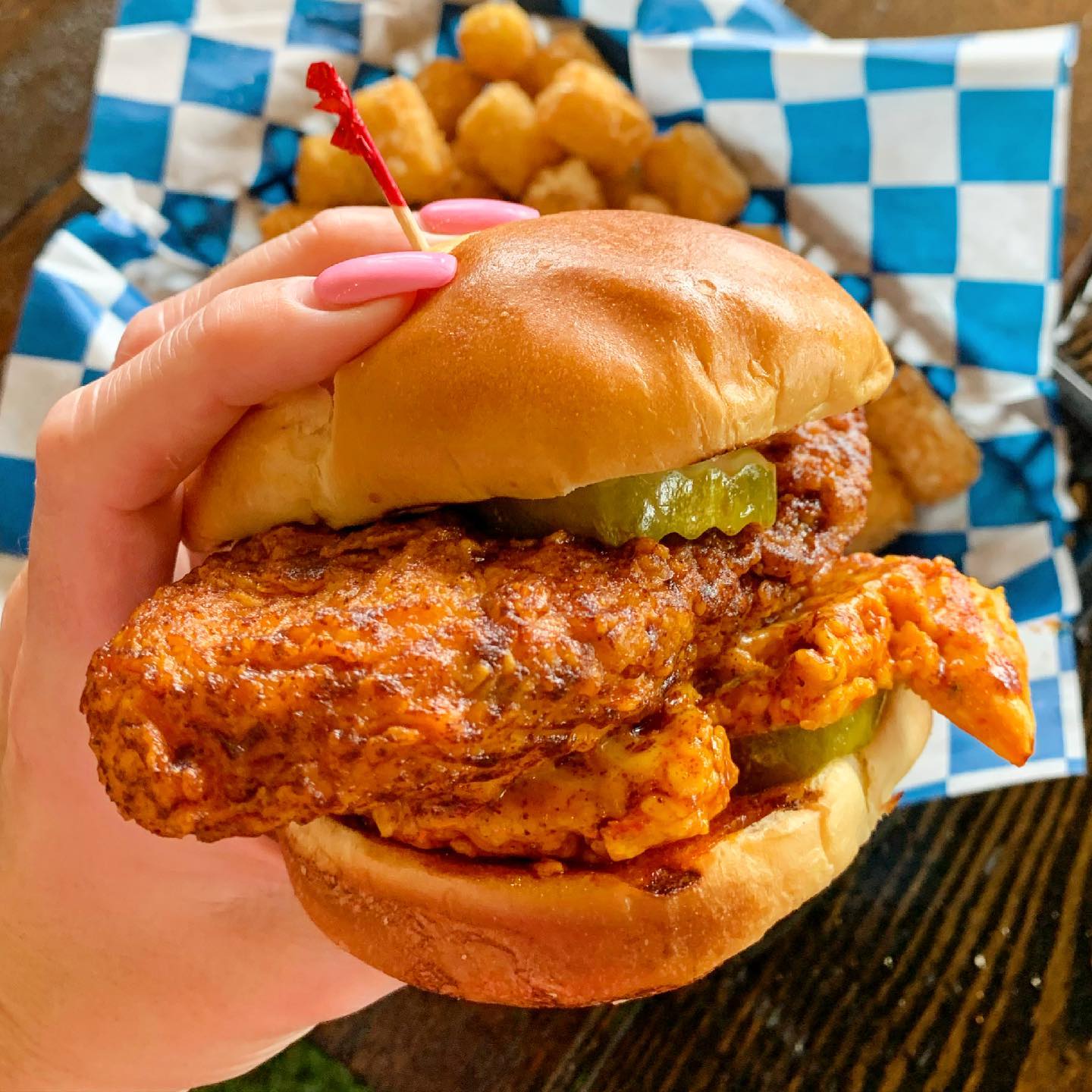 a manicured hand holding a hot chicken sandwich in front of a pile of tater tots