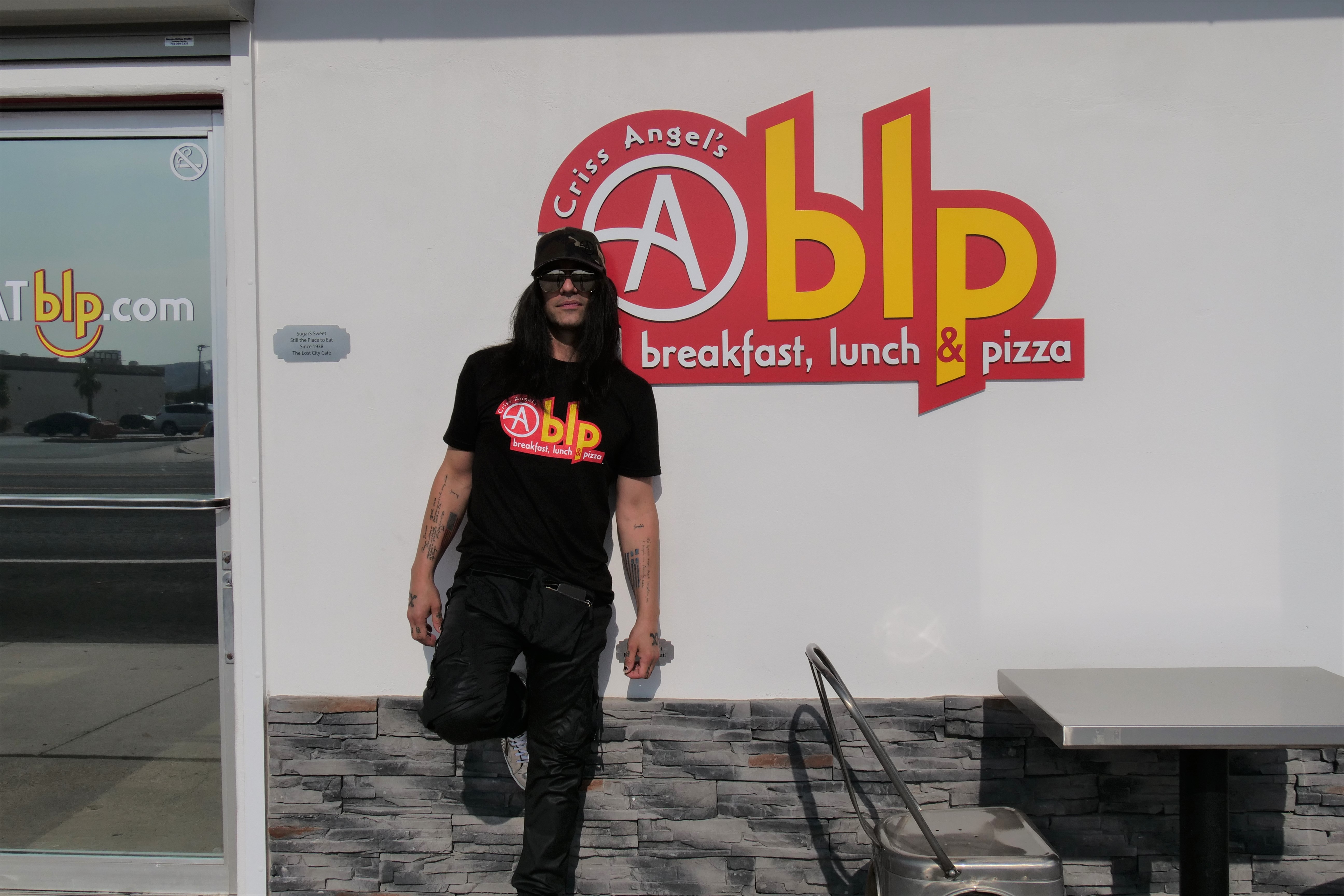  A man in a black hat, shirt, and pants with sunglasses on stands in front of a sign that says Cablp