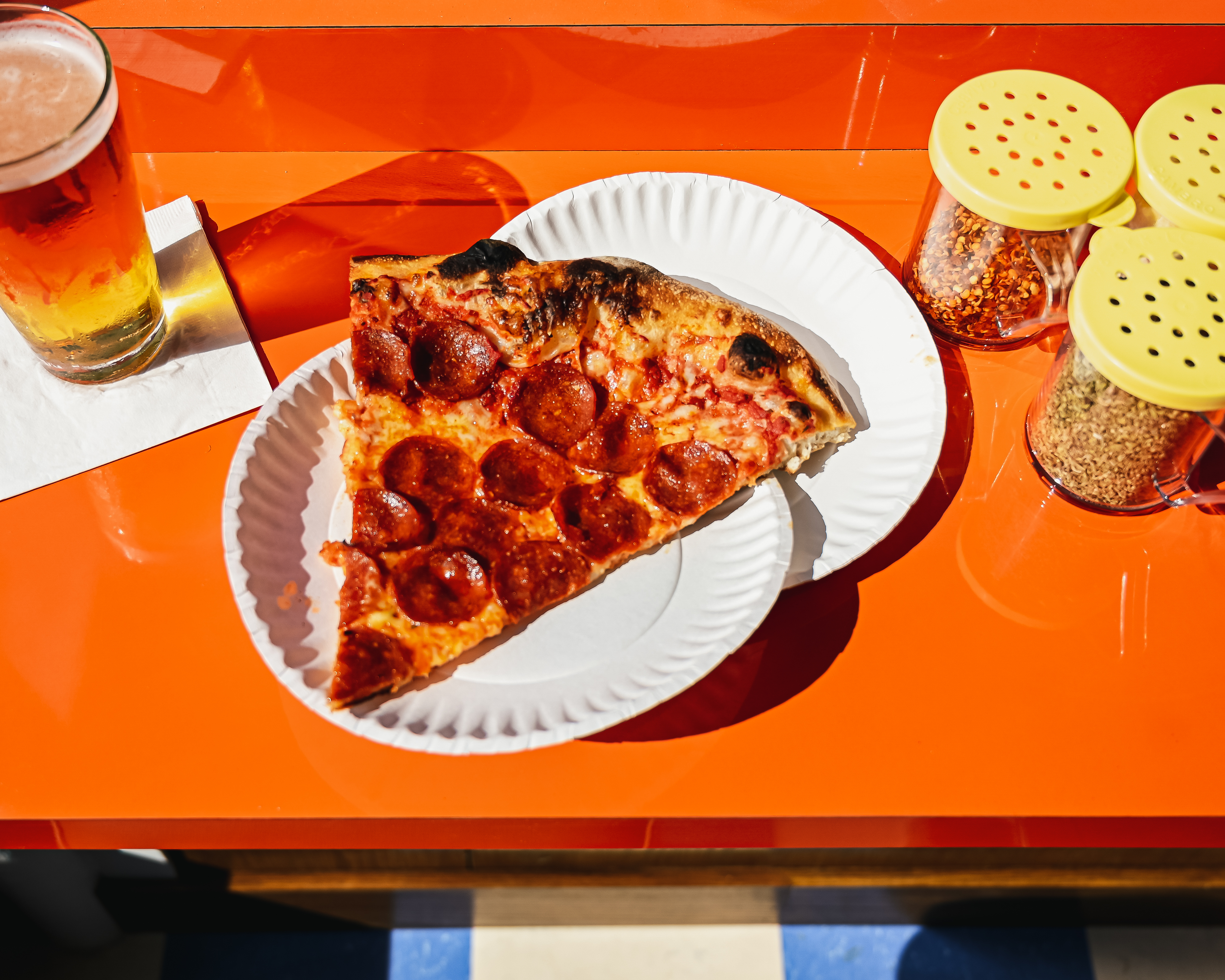 A pepperoni pizza slice on two white paper plates on an orange table with a glass of beer to the left and pizza topping containers to the right