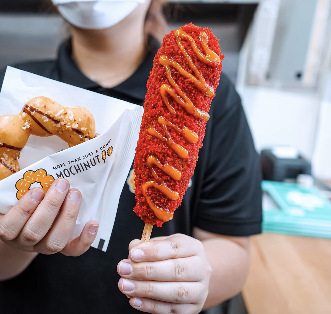 A person holds a mochi doughnut and a corn dog on a stick covered in bright red powder