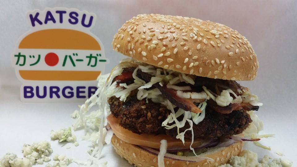 A giant fried patty burger stuffed with toppings such as shredded lettuce, with the Katsu Burger logo in the background against a white backdrop.