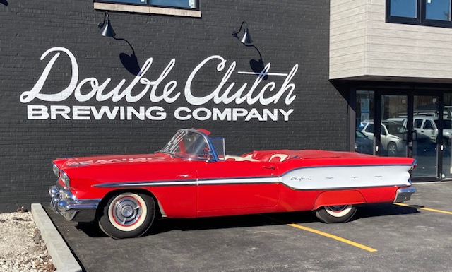A bright red vintage convertible with white fins parked beside a black building with the words “Double Clutch” painted in white