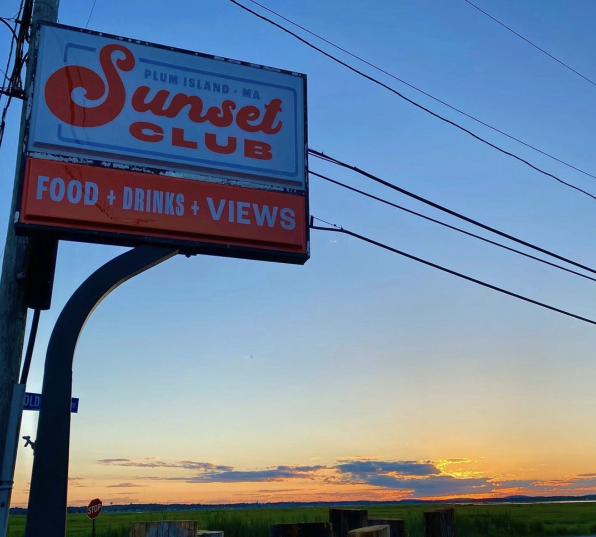A white sign with orange text that reads “Sunset Club: Food + Drinks + Views” backdropped by a sunset over a marsh