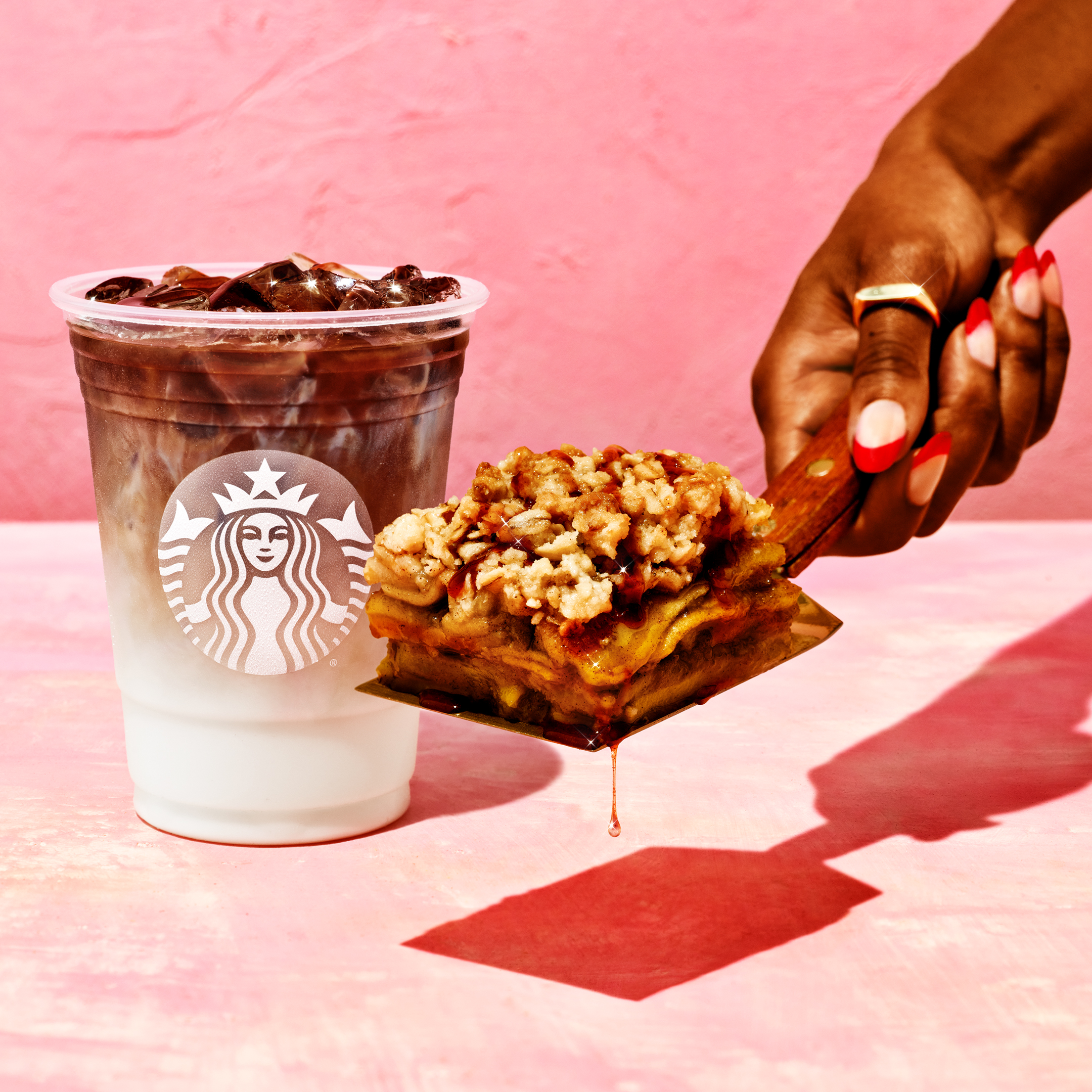 A plastic cup with a white and brown layered starbucks drink in the background. In the foreground, a manicured hand holding a spatula with a slice of crisp