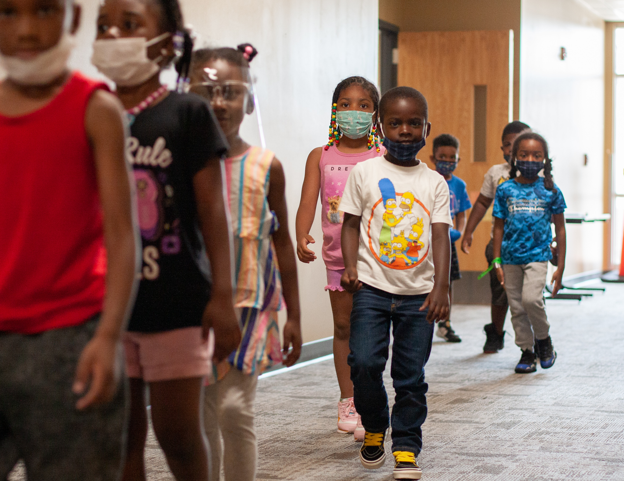 Eight kids, all wearing masks or face shields, walk in a jumbled line through a carpeted school hallway