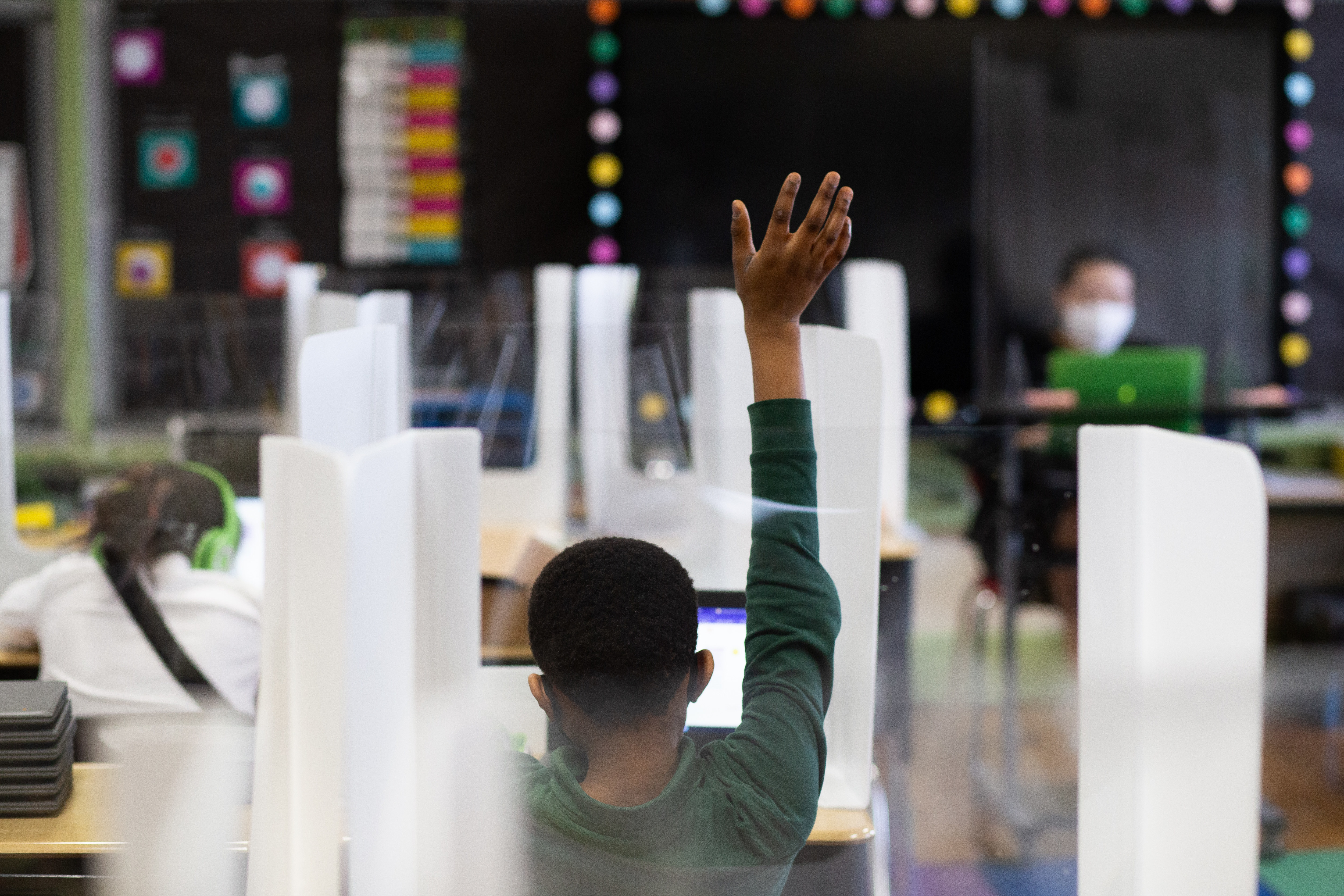A fourth grade student raises his hand in class. The photo shows the back of the student’s body with his hand in the air. The teacher is visible in the background of the photo, facing the camera. The teacher is wearing a face mask. The student’s desk is surrounded by plastic dividers.