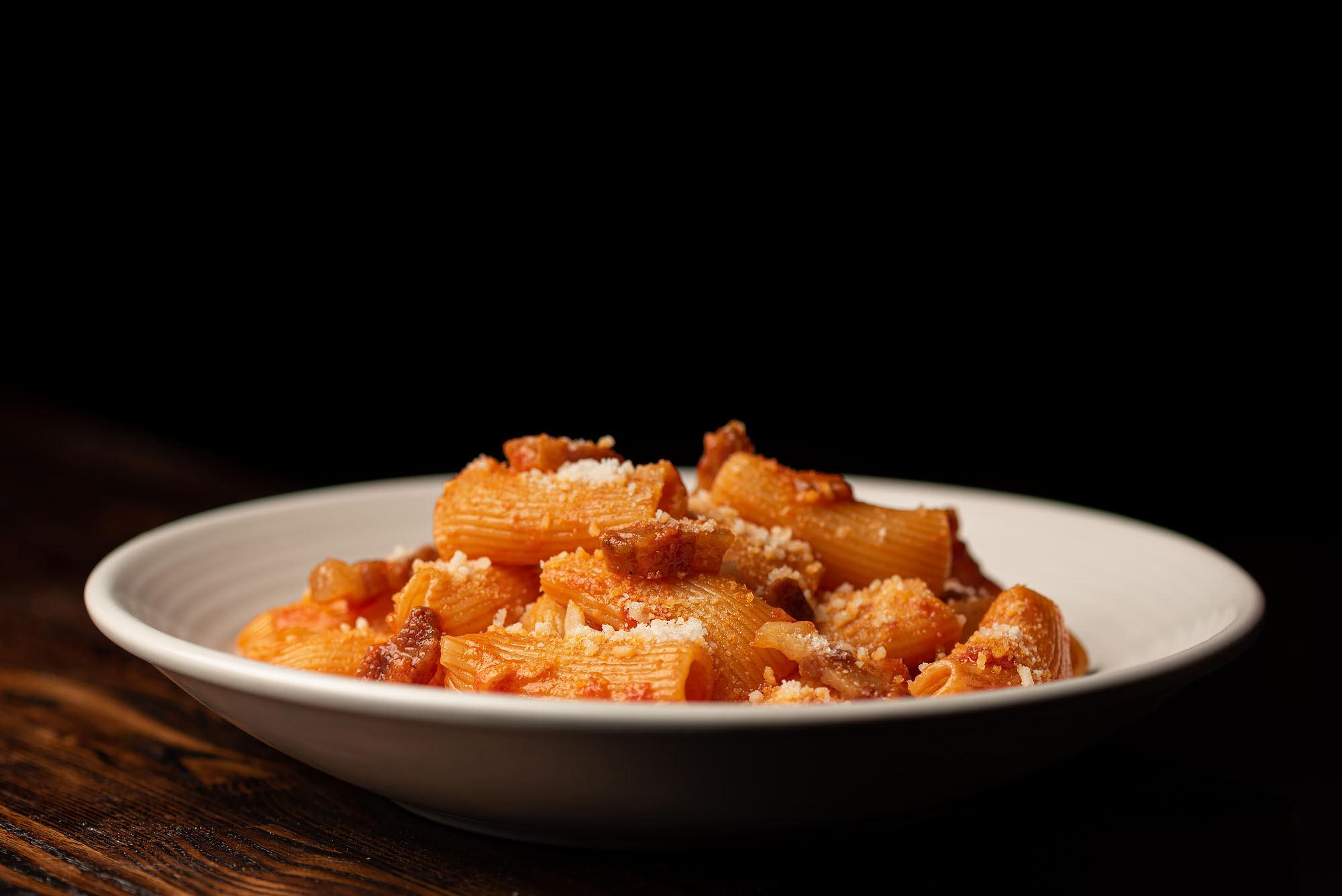 Rigatoni amatriciana at Fingers Crossed, shown in a dark room and from the side, inside a round white plate.