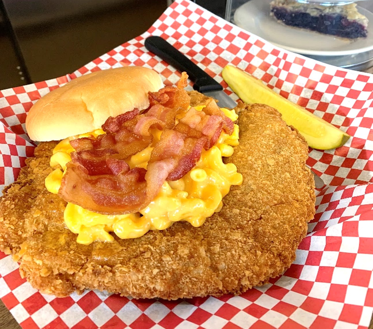 A huge fried pork tenderloin topped with mac and cheese, bacon, and a top bun, sitting in a checkerboard paper-lined basket