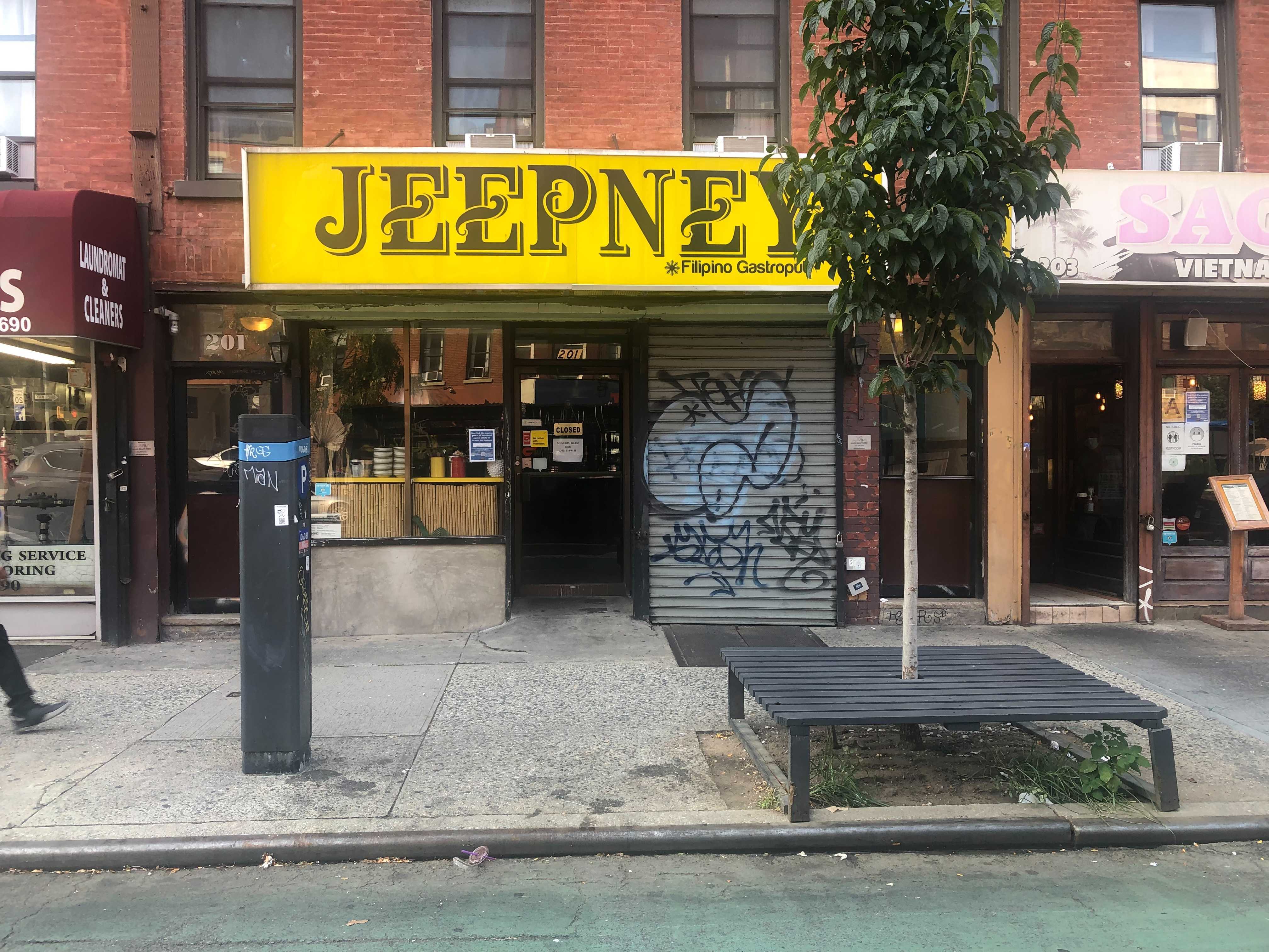 A New York City storefront with a closed steel grate that is covered in graffiti. The restaurant’s yellow sign reads “Jeepney” in brown lettering.