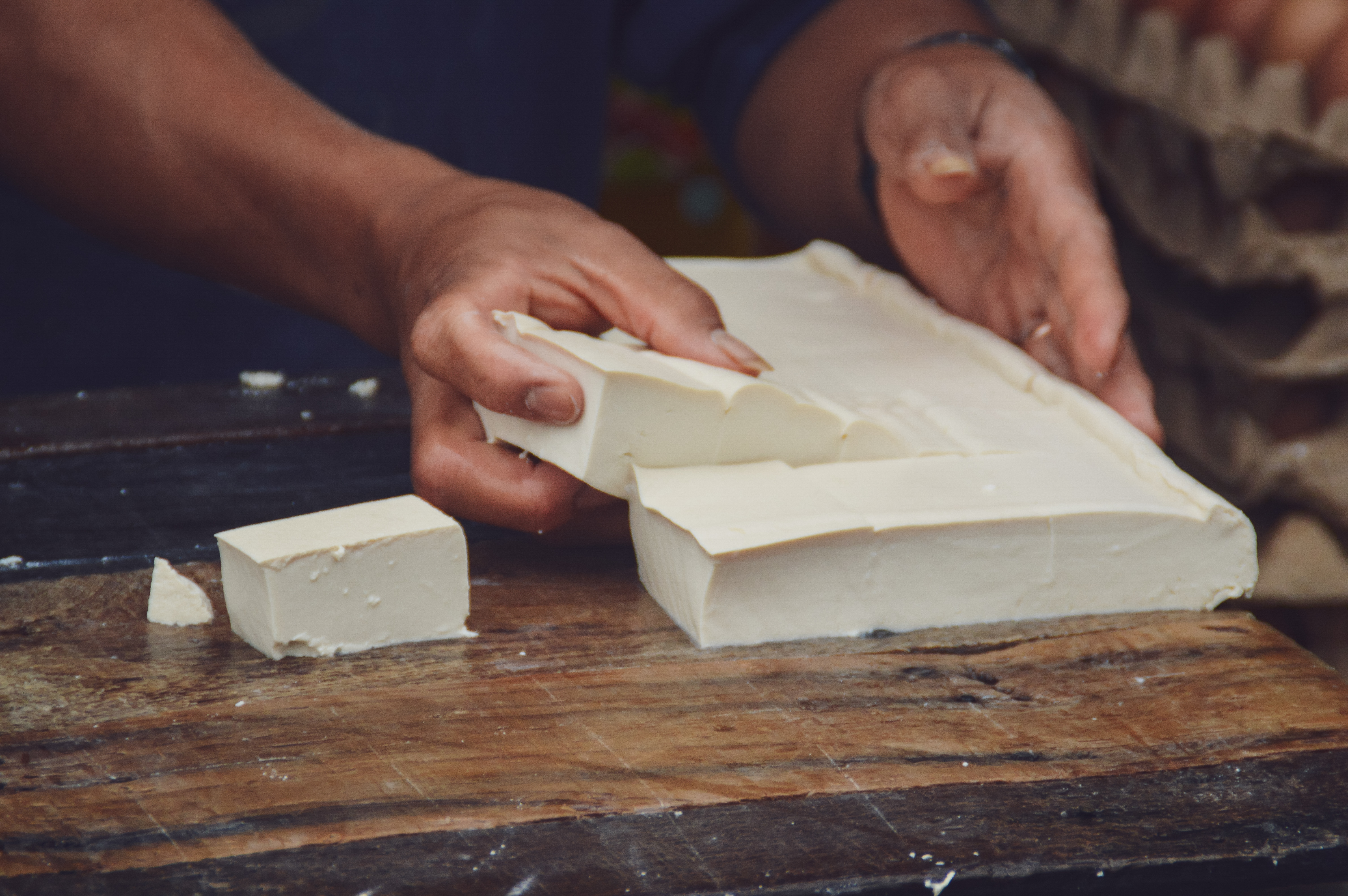 A close-up of a person’s hands as they work a fresh brick of tofu on a wooden table.
