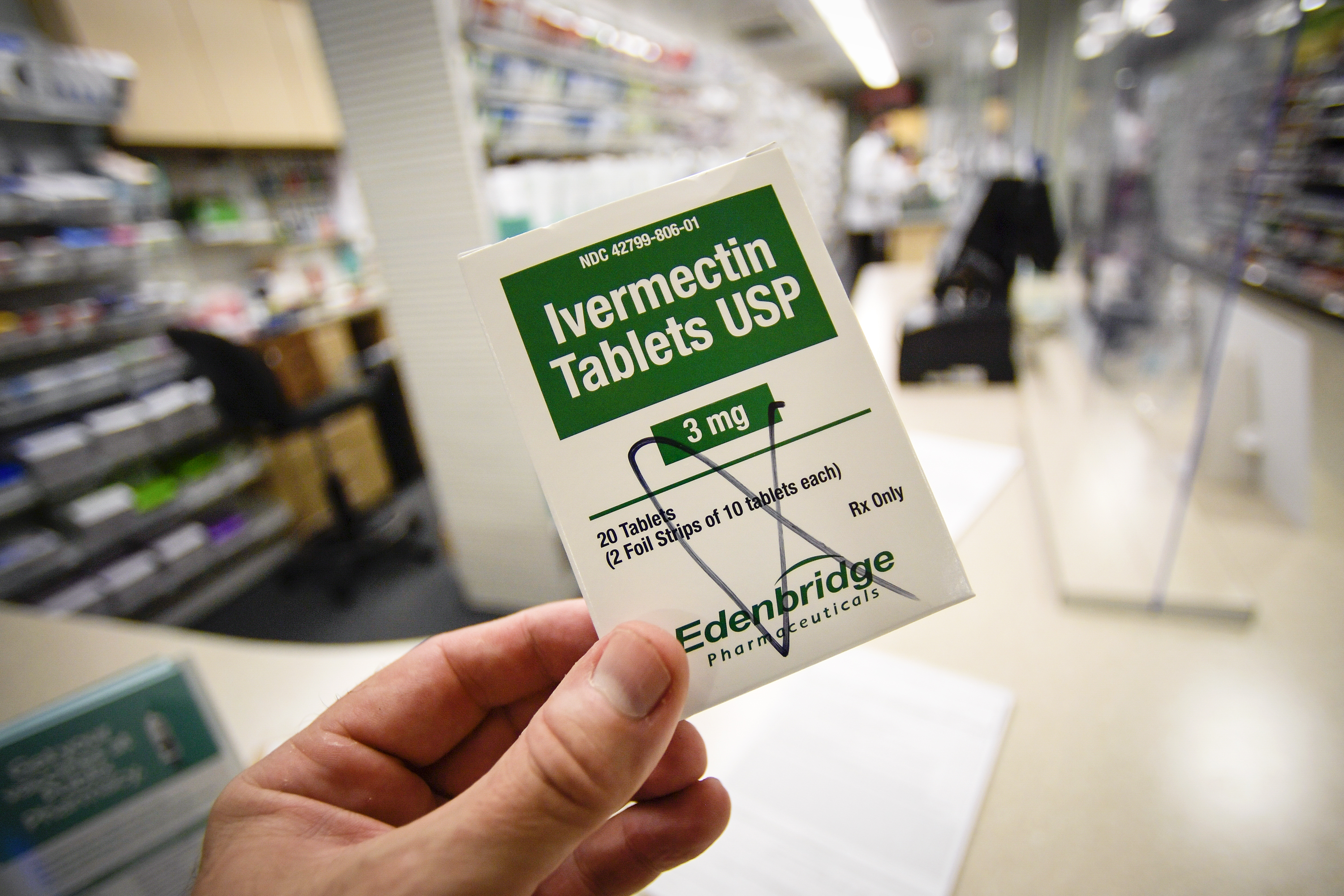 A hand holding a small packet labeled Ivermectin Tablets USP. In the background is the aisle of a drugstore.