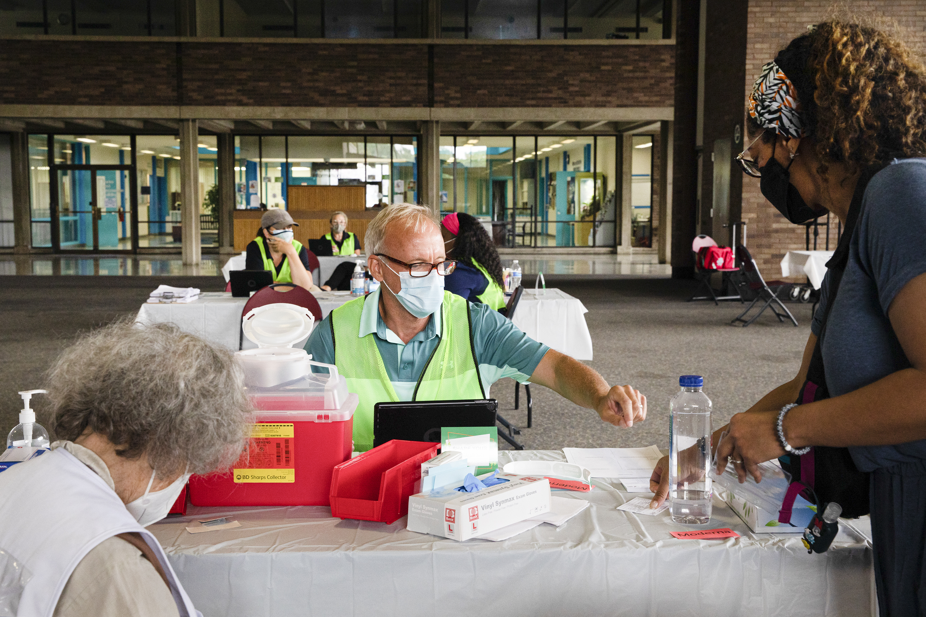 A patient shows their vaccination card to a person sitting at an outdoor table distributing vaccine shots.