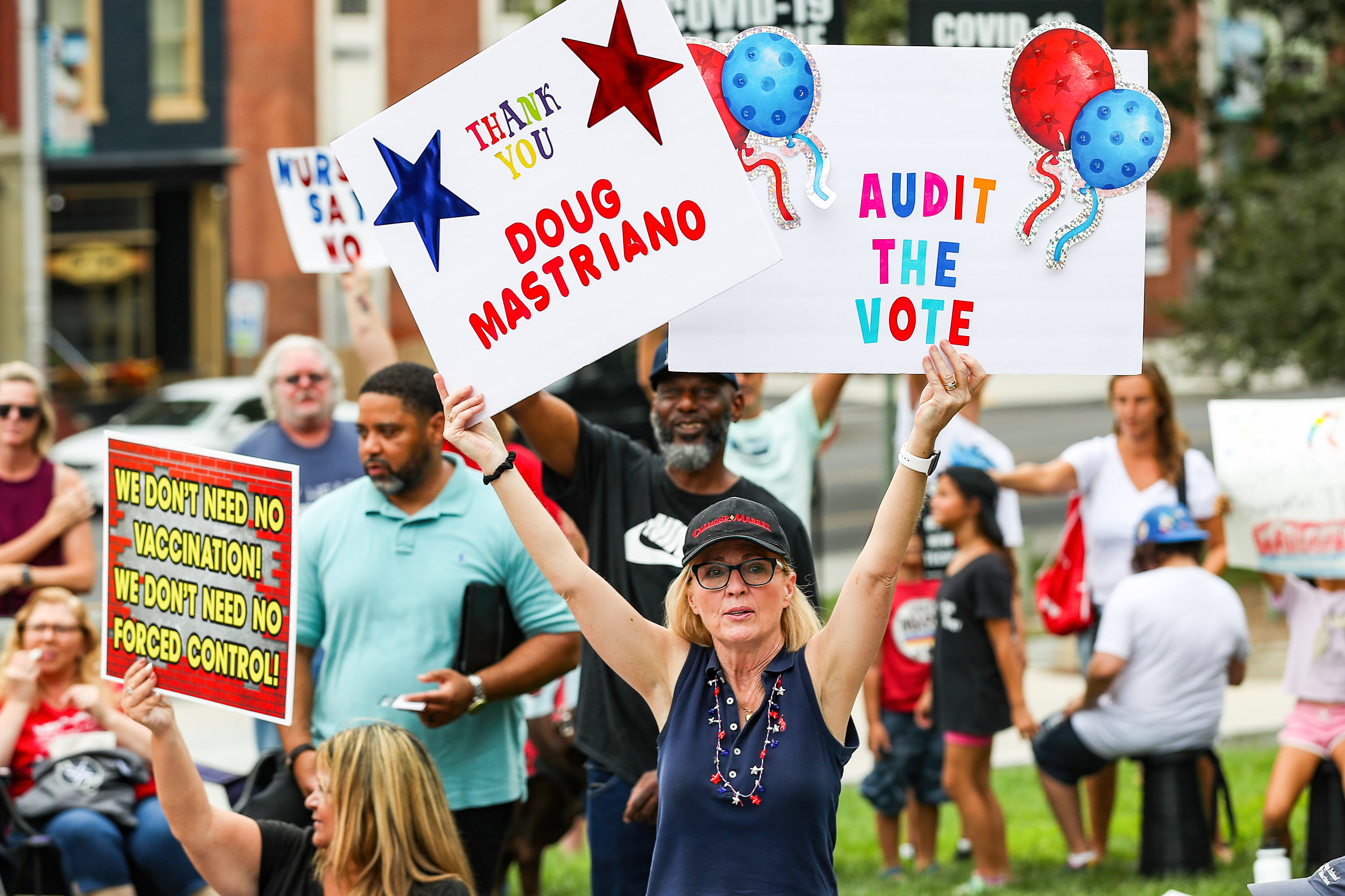 A woman amid a crowd of protesters holds up two colorful signs, one reading “Audit the vote” and another reading “Thank you Doug Mastriano.”