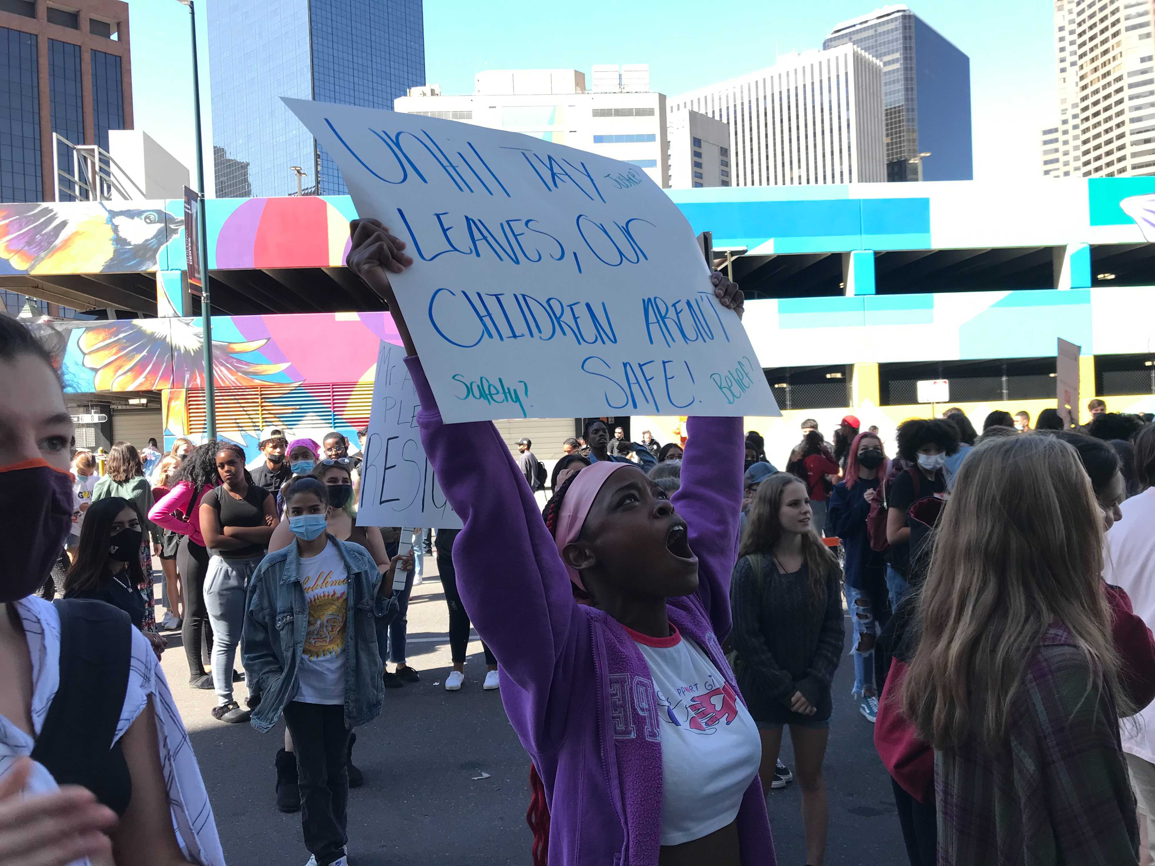 A high school student wearing a purple sweatshirt stands in a crowd. Her arms are raised, holding a sign that reads “Until Tay leaves, our children aren’t safe.” She is shouting.