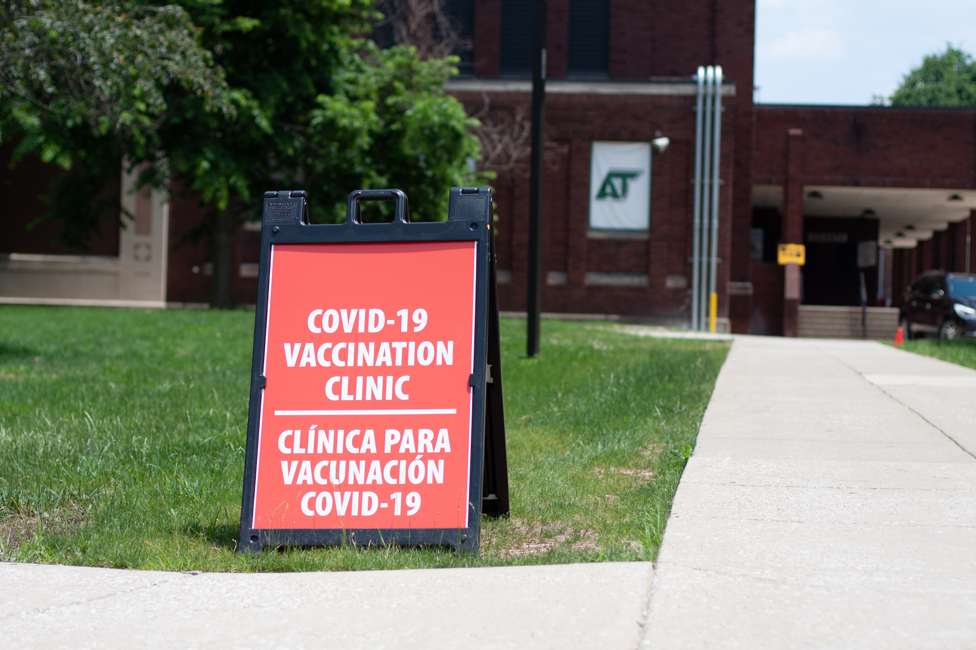A red clapboard sign sits on a patch of green grass in front of a red brick school building. The sign reads “covid-19 vaccination clinic, clínica para vacunación de covid-19”