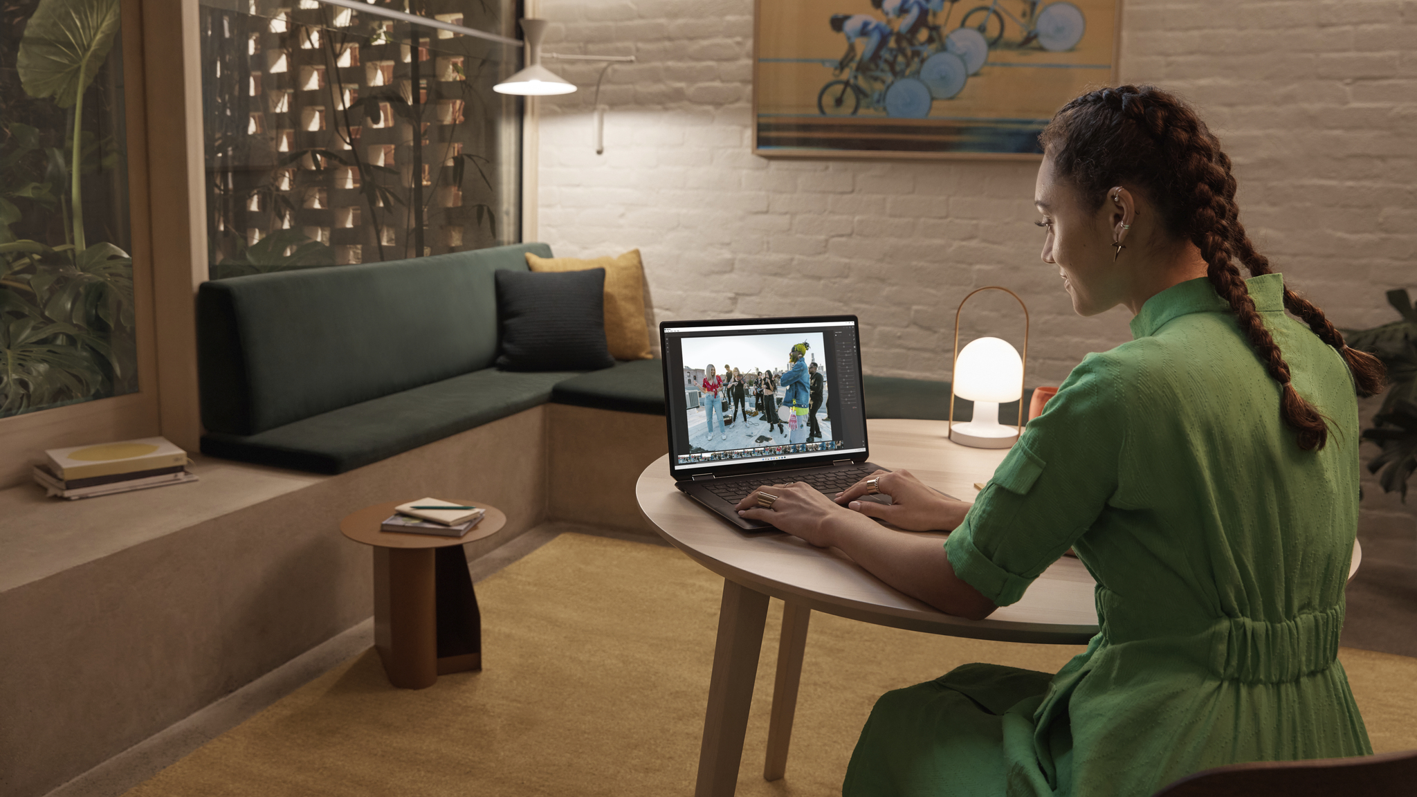 A user in a red dress types on the HP Spectre x360 16 in an evening room at a small wooden table. The screen displays a photo of a group of urban dancers in an outdoor scene in an editing program. To its left is a small lit lamp.