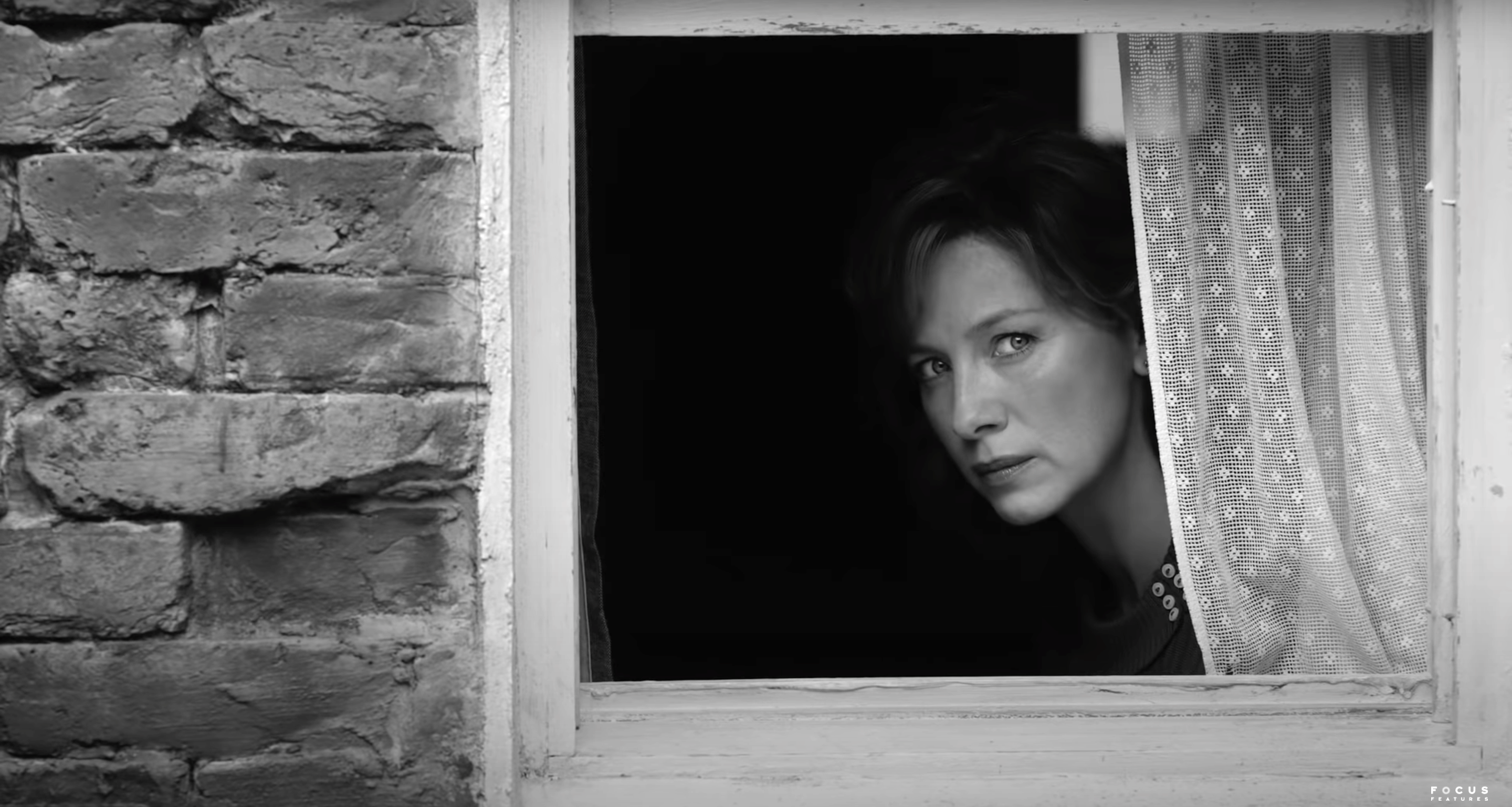 A woman peeks from behind a window in a black and white still image.