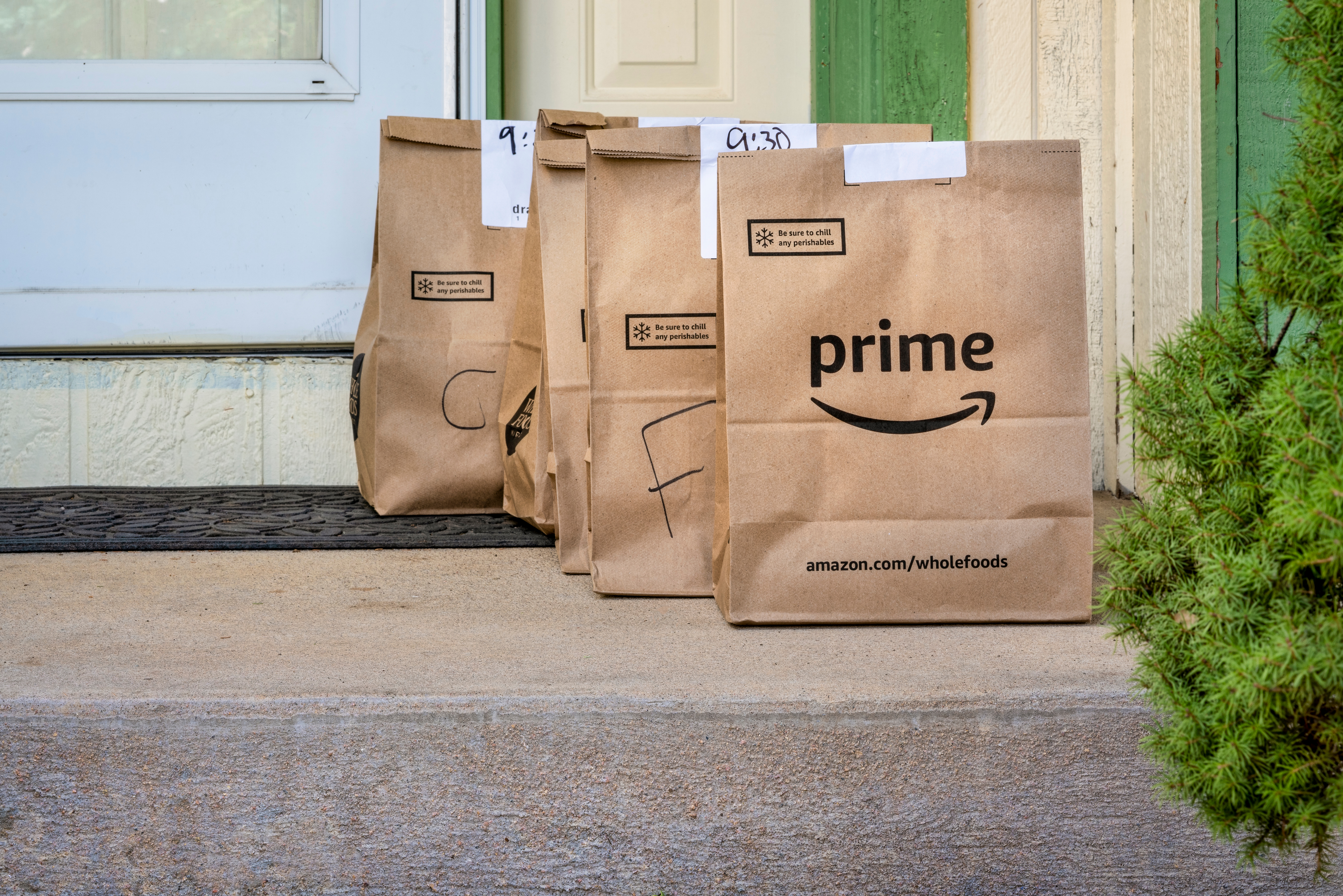 Three brown paper Amazon Prime Whole Foods delivery bags sit on a front stoop, with the corner of a white front door visible in background.