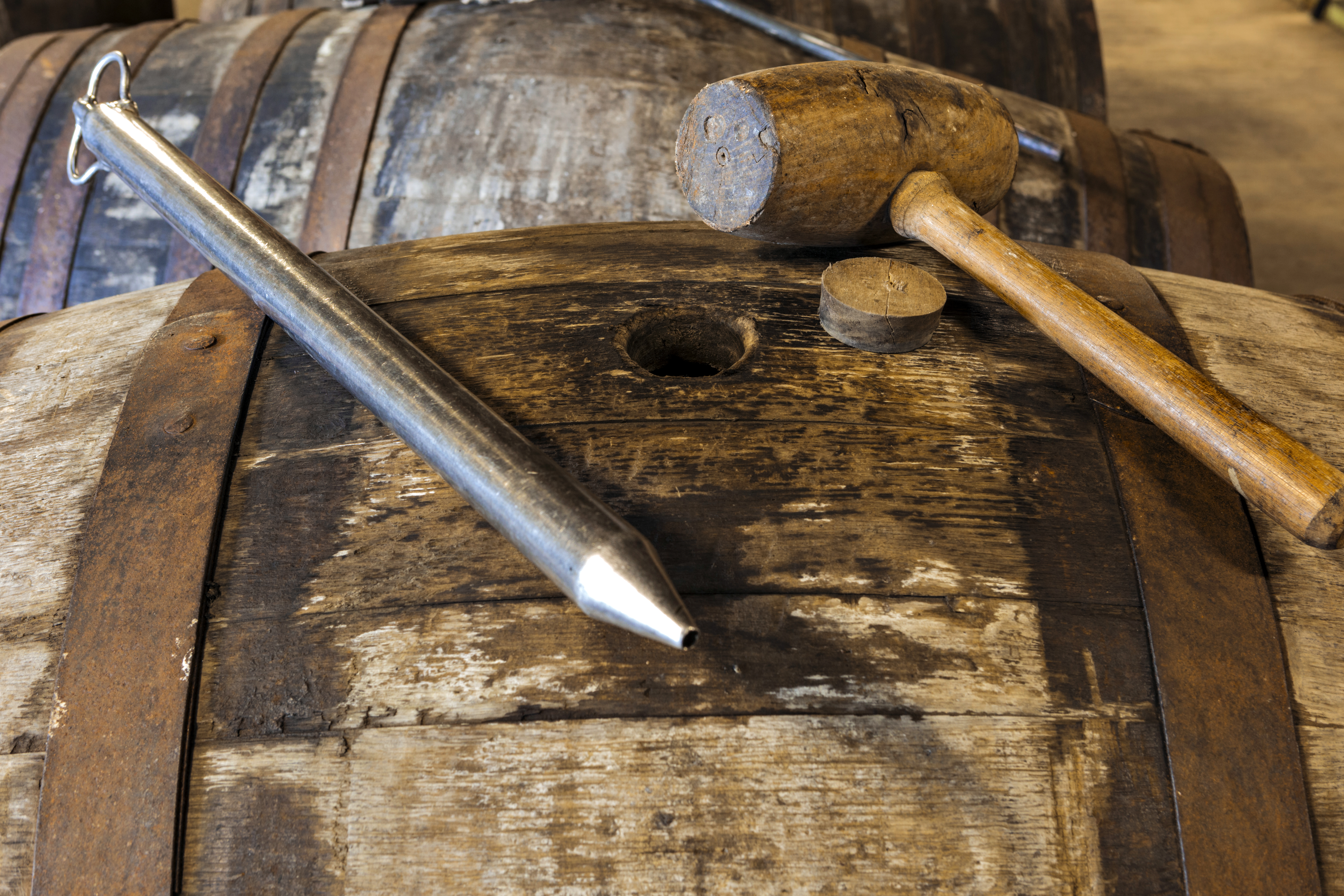 A charred barrel lies on its side with its round opening facing up. Resting on the barrel is a long metal pipette, a cork, and a wooden mallet.