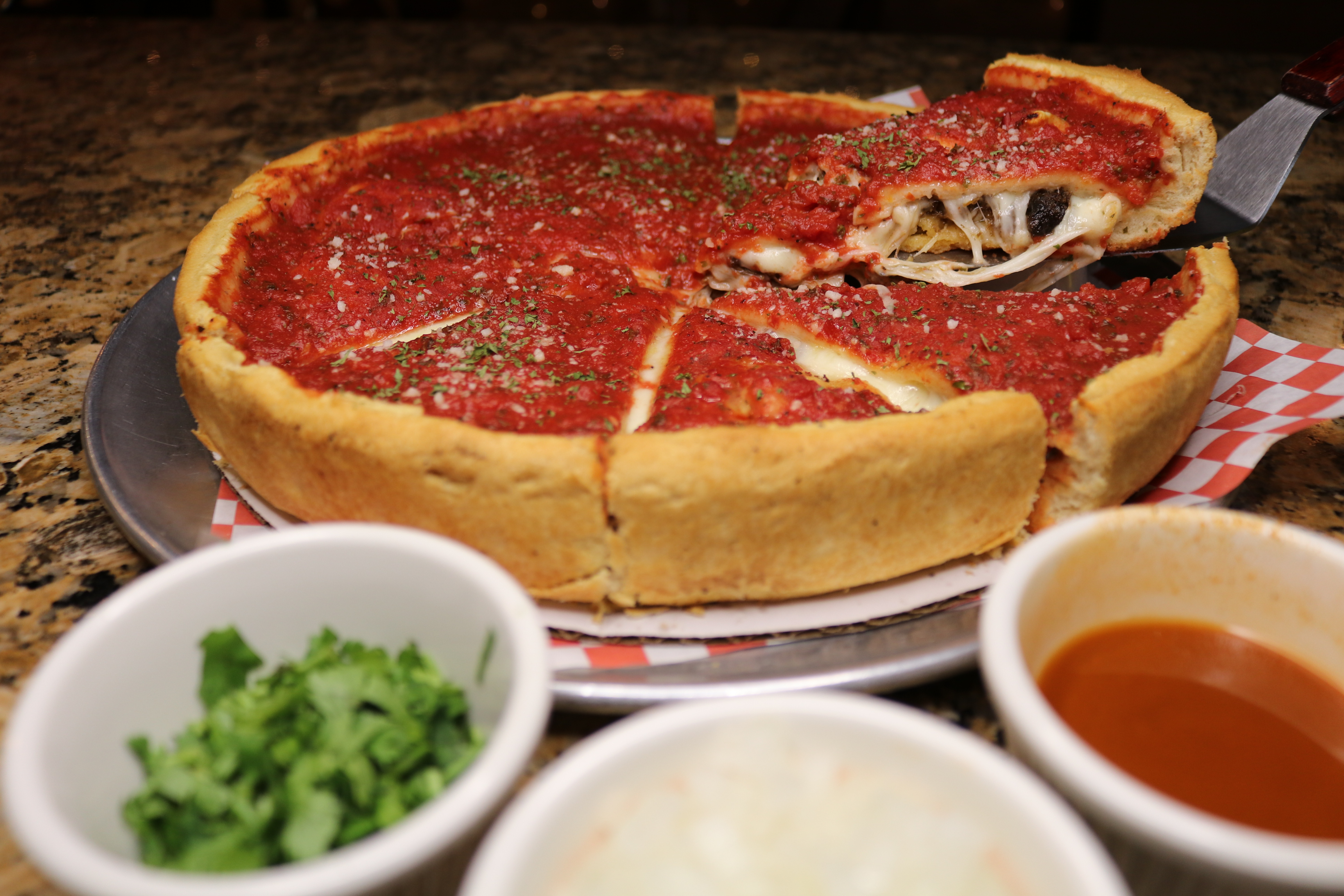 A spatula lifts up a single wedge from a deep dish pizza; bowls of cilantro, onions, and consomme in the foreground