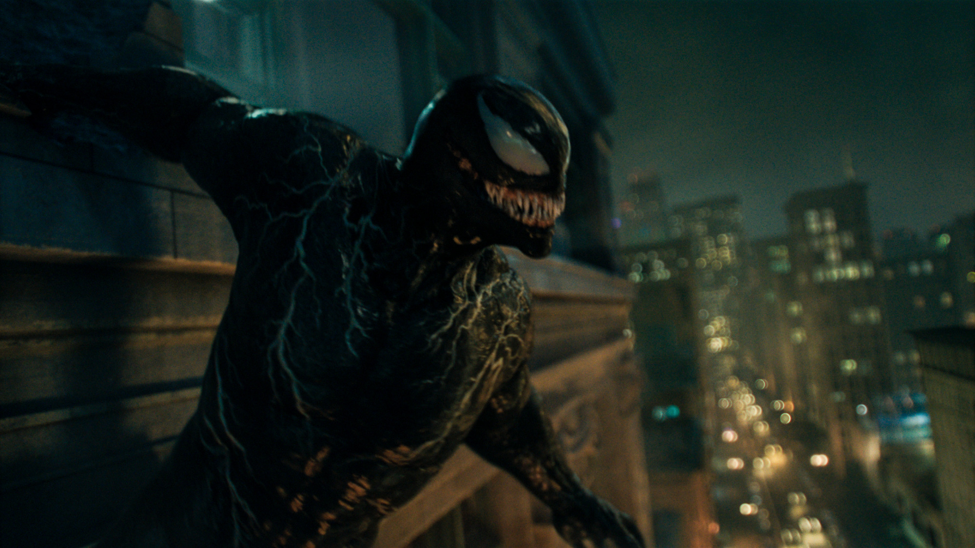 Venom perched on the side of a tall building at night with a skyline and lights below him.