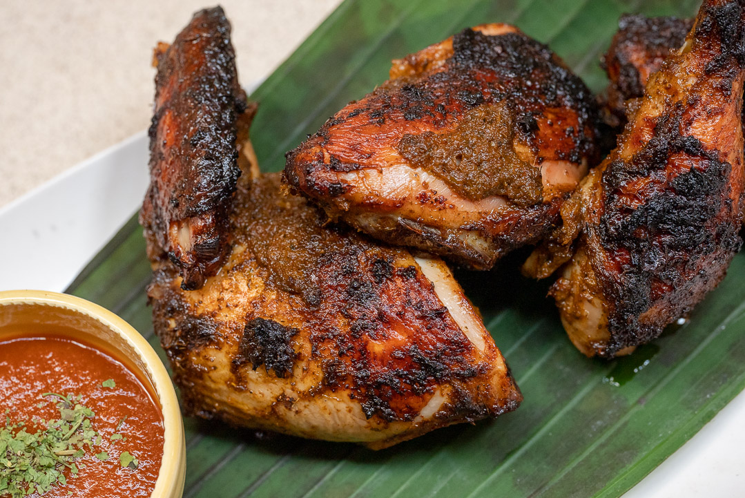Three pieces of dark-roasted chicken on a large green leaf next to a bowl of reddish sauce with green sprinkled leaves.