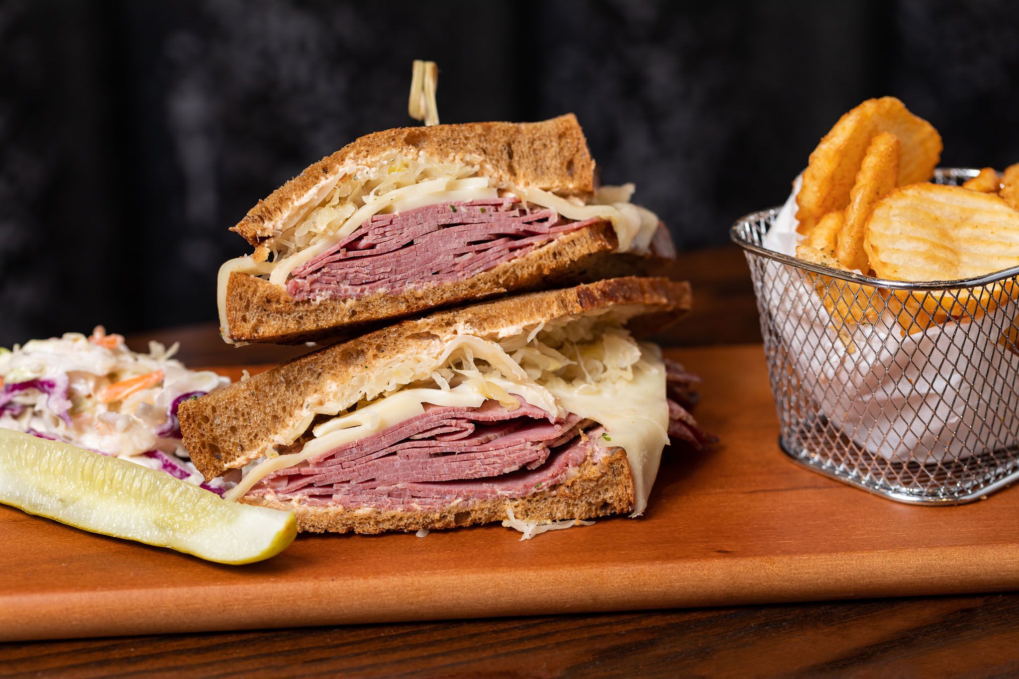 a rueben sandwich and cup of fried potatoes on a wooden bar