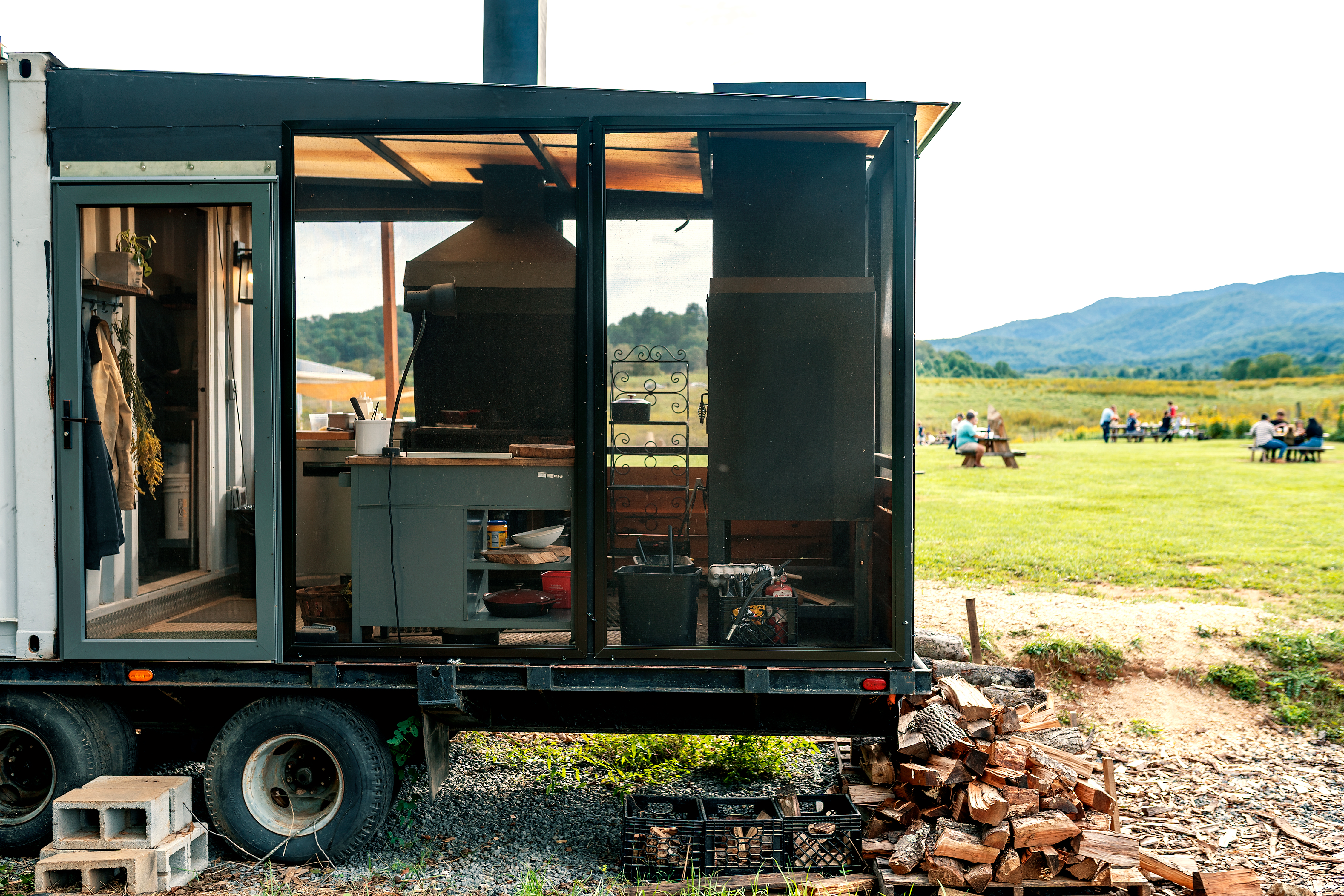 A view of two black, custom wood-burning hearths through the open, framed walls of a trailer extension that looks out over a field with a mountainous backdrop.