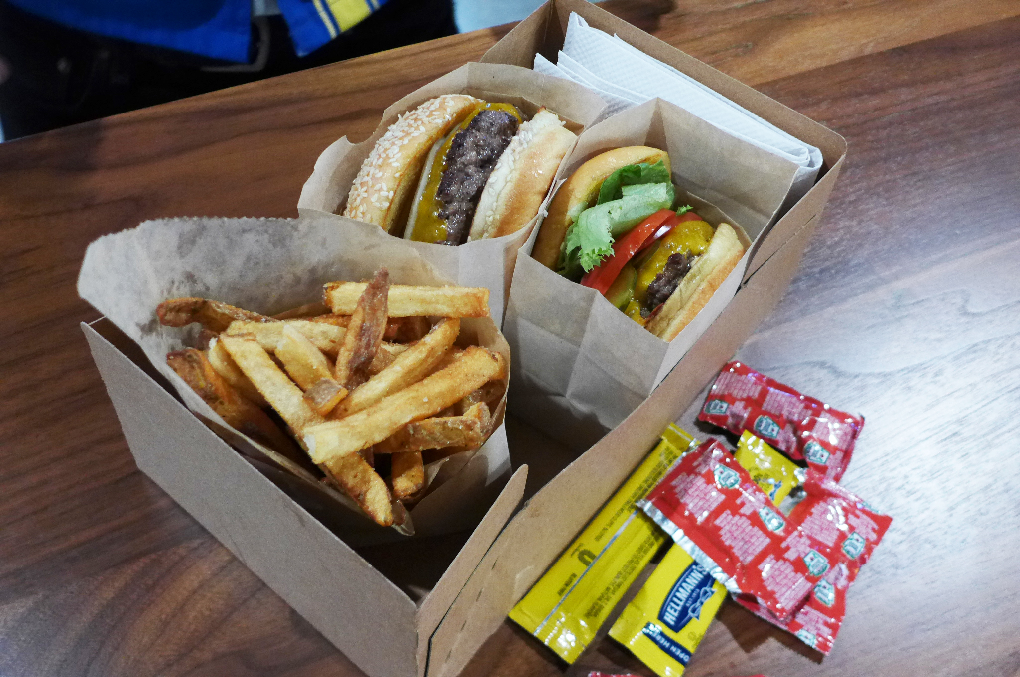 Two burgers in boxes, one with salad stuff on it the other without, on sesame seed buns with french fries on the side.