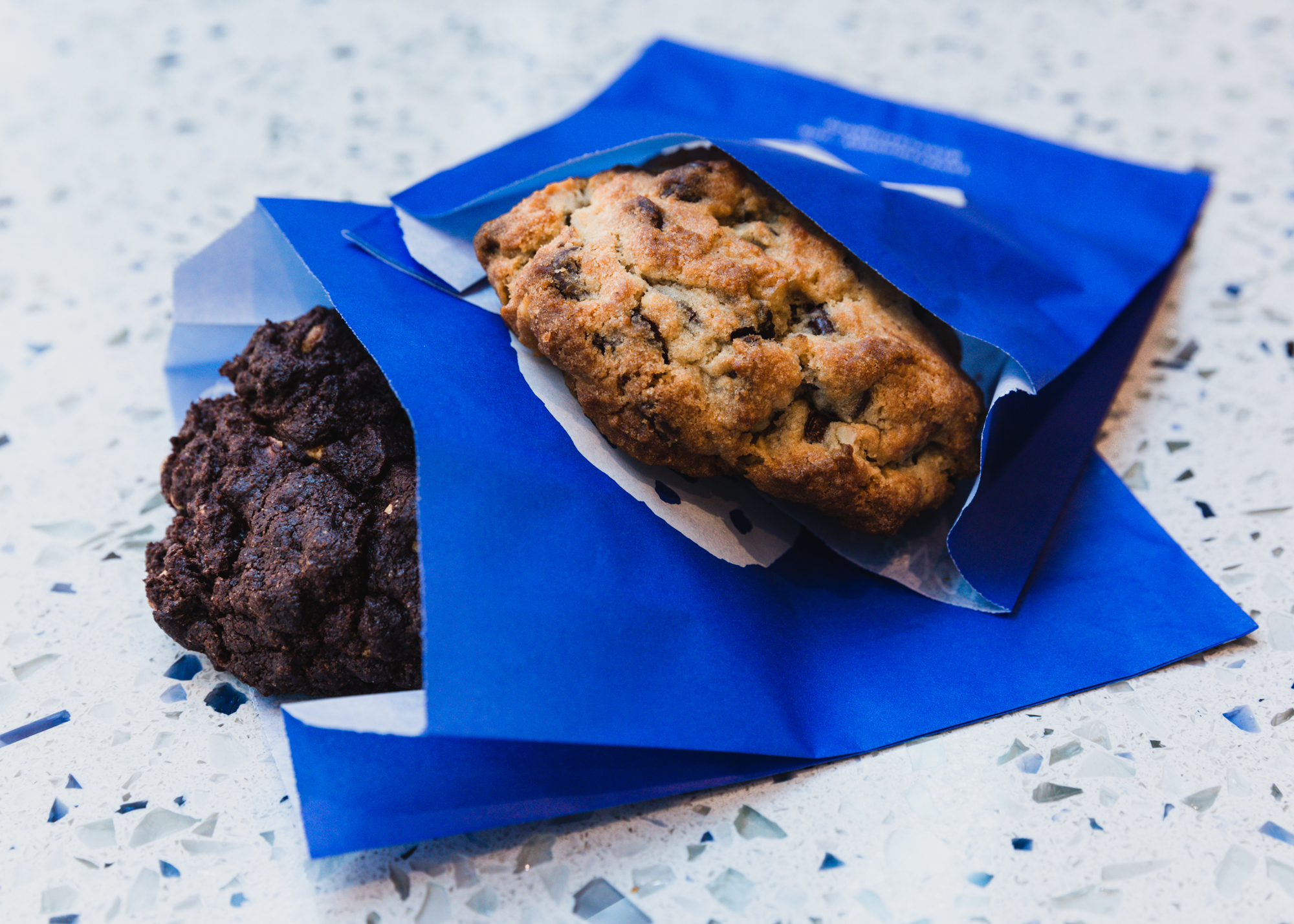 Two cookies —&nbsp;one chocolate chip, one dark chocolate —&nbsp;are in individual bright blue paper sleeves, sitting on a countertop.