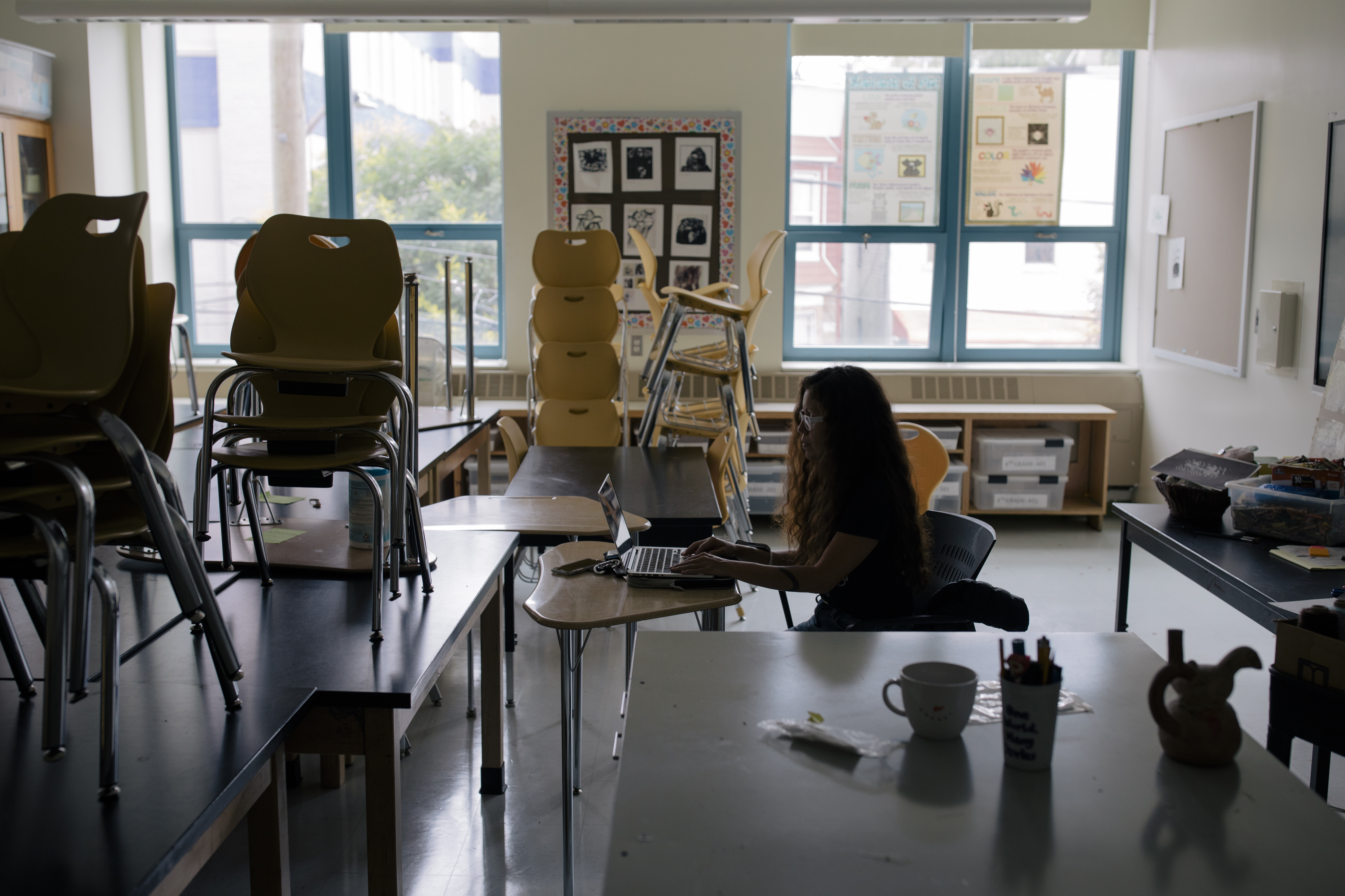 Art teacher Elizabeth Velasquez begins a Zoom class in her empty classroom. The chairs are stacked neatly on desks as she sits at a small desk in the front of the dark classroom.