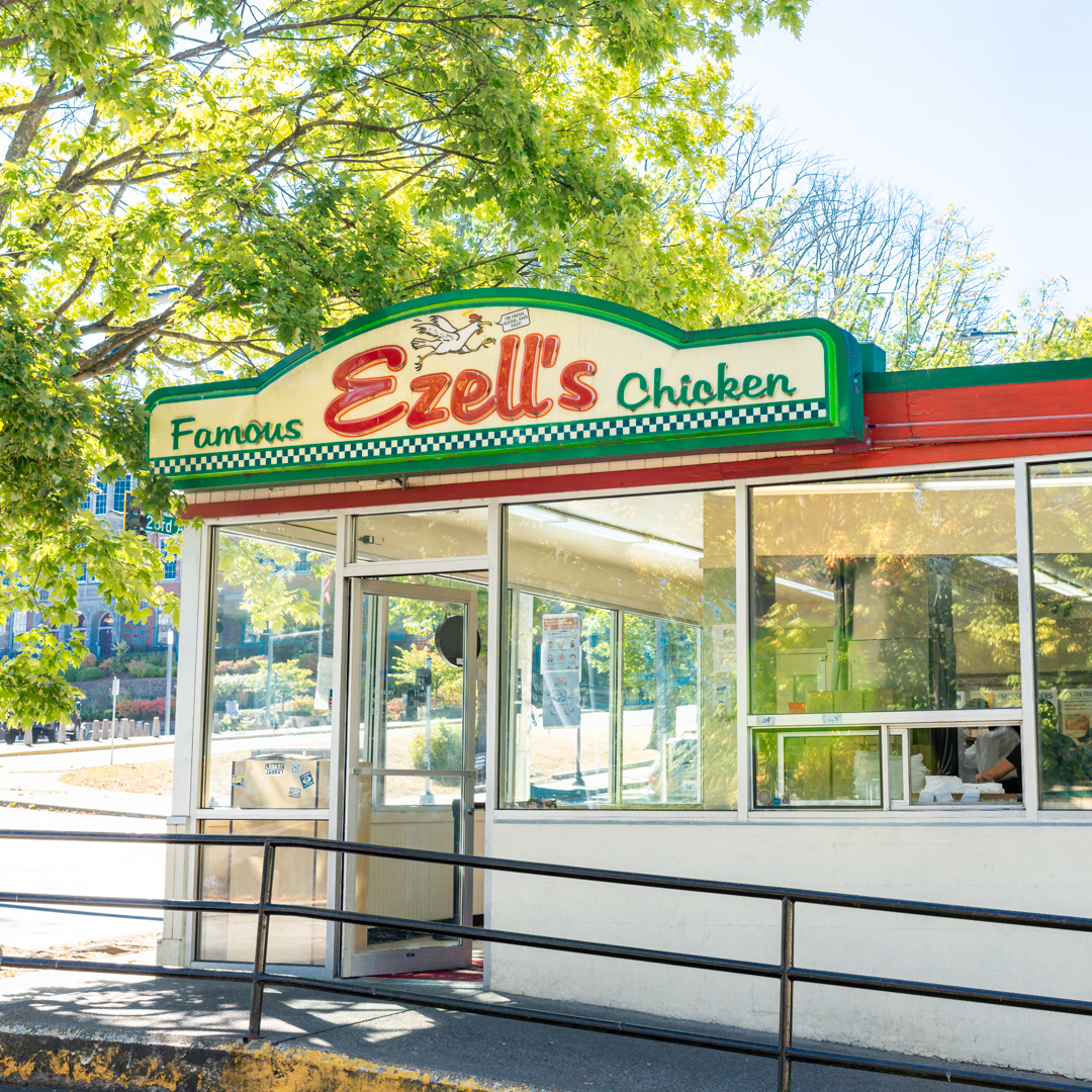 The exterior of Ezell’s Famous Chicken restaurant, with glass windows, and a sign with green and red lettering and a cartoon chicken.