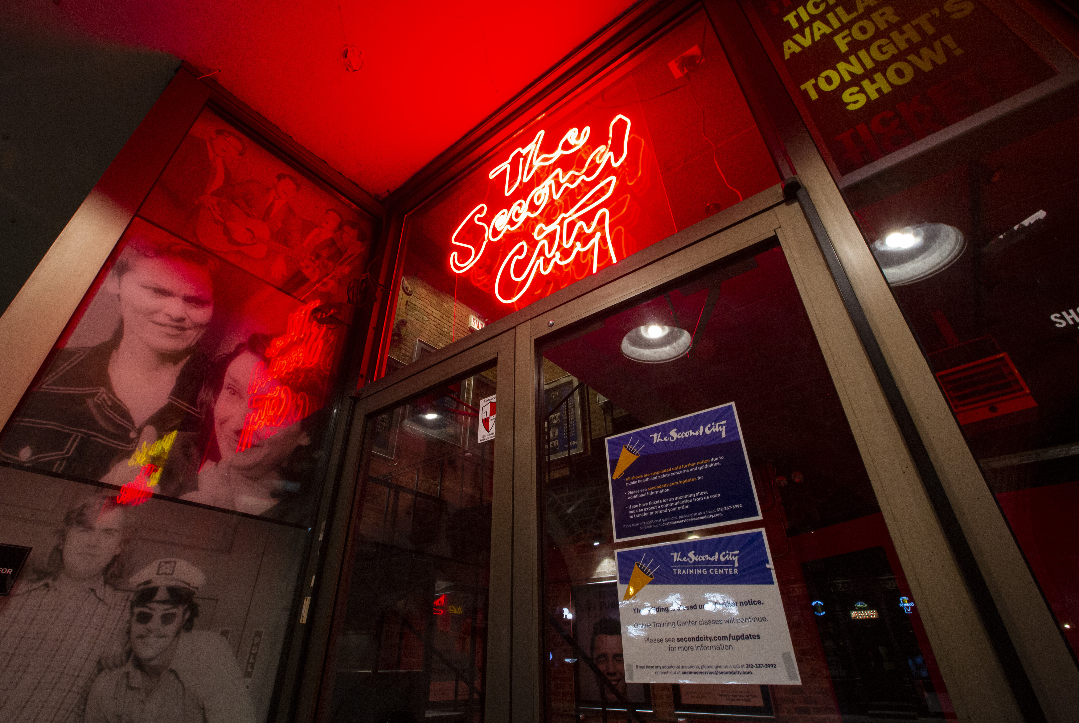 A glass door with posters to the left and a red neon sign that says “The Second City” above