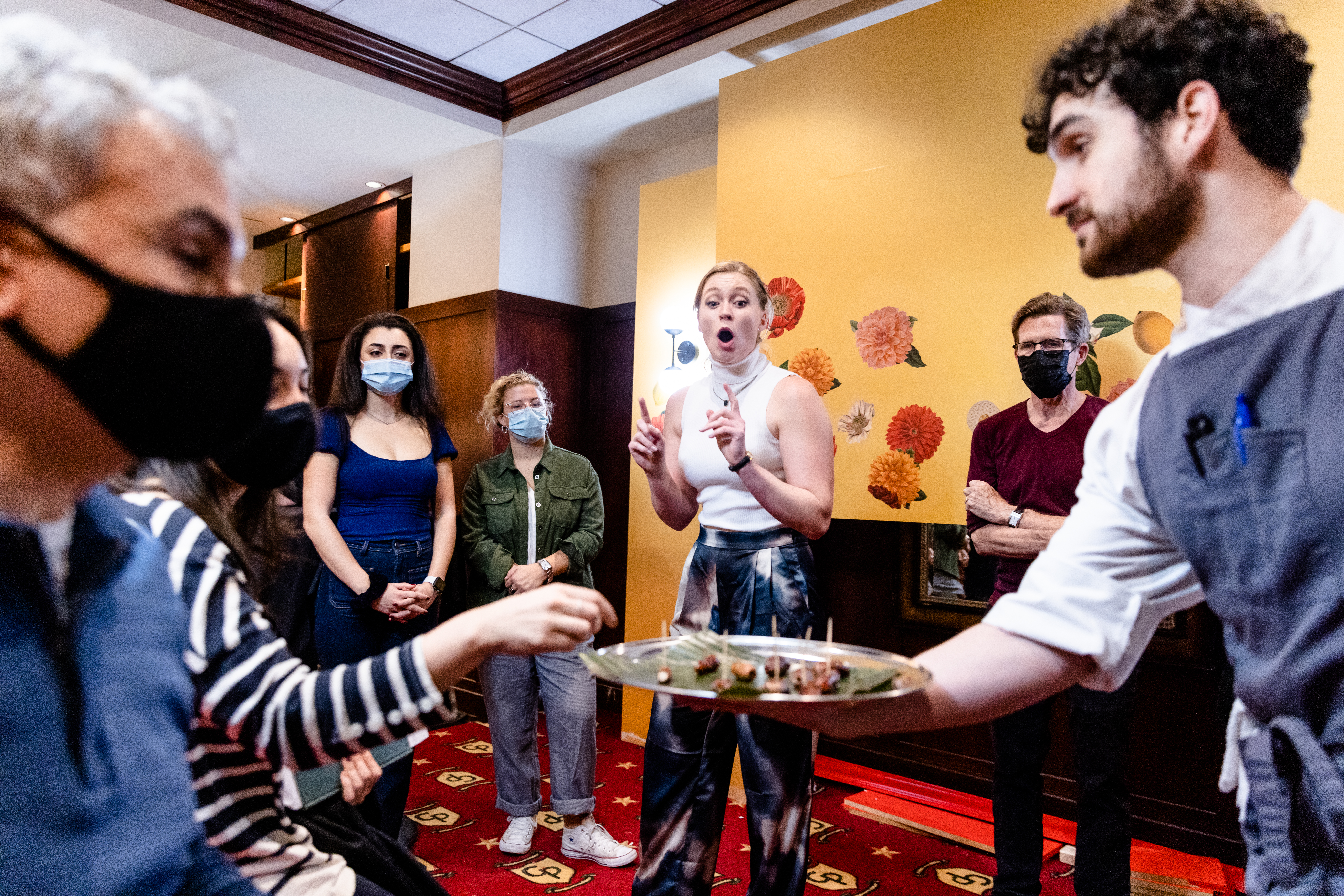 A waiter handing a platter of food to a masked person sitting down while a woman gasps in shock.
