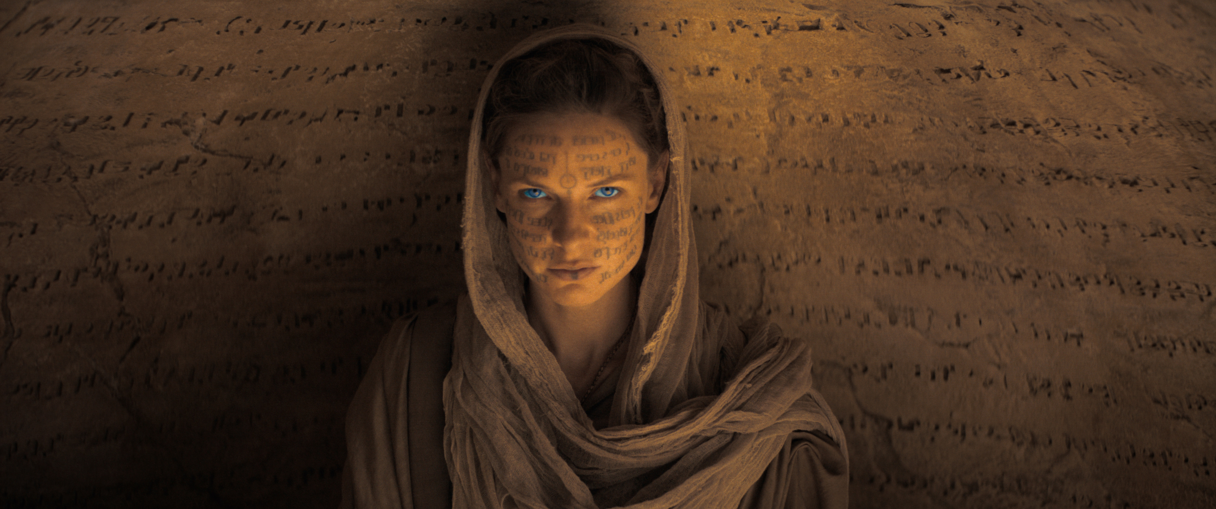 Rebecca Ferguson, in sandy robes, sits in front of a wall carved with lines of alien text. Her face is also scrawled with alien text, and her eyes are glowing blue in Dune.