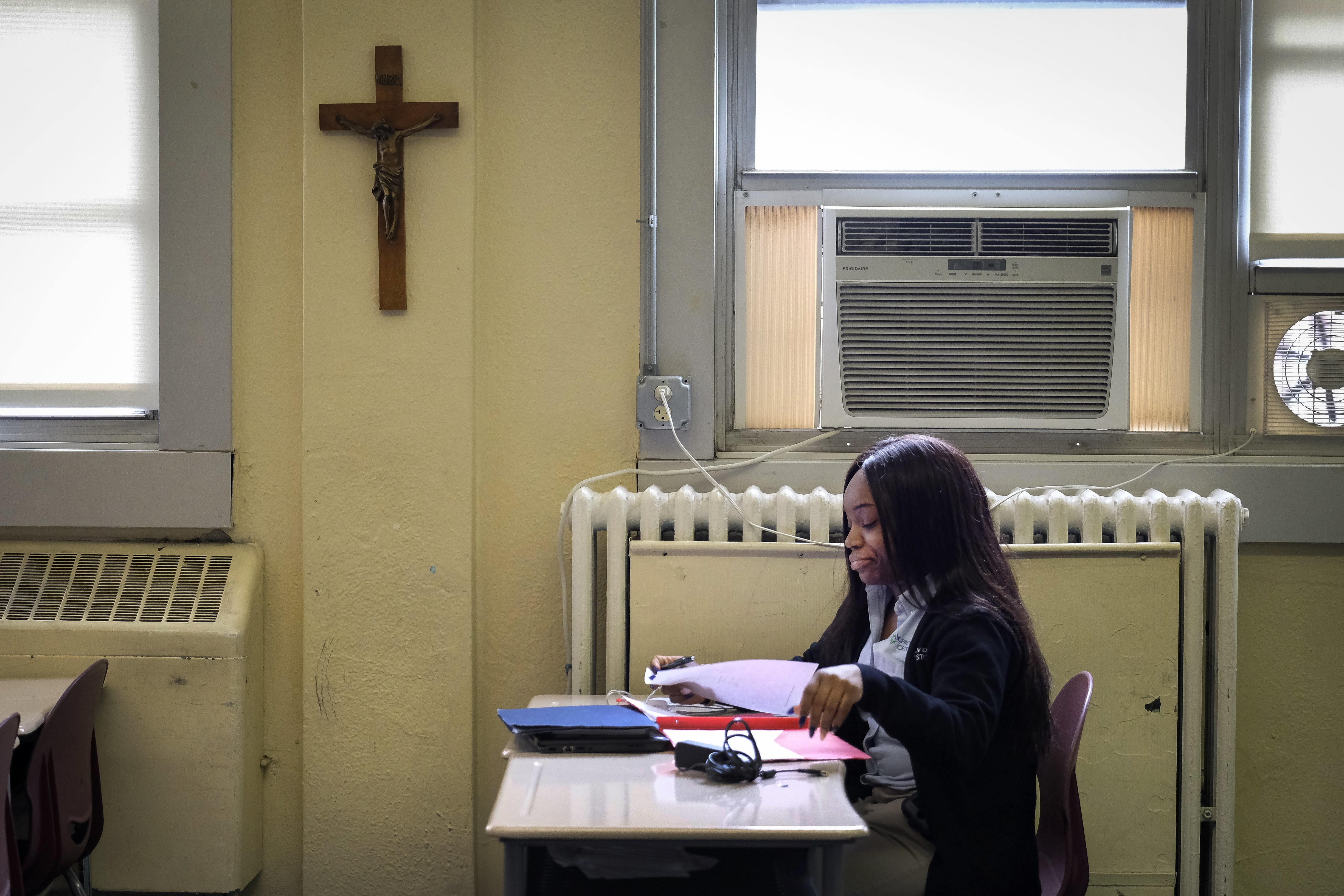 A cross hangs on a wall near a student looking through papers at a desk in a classroom in a Catholic school. 