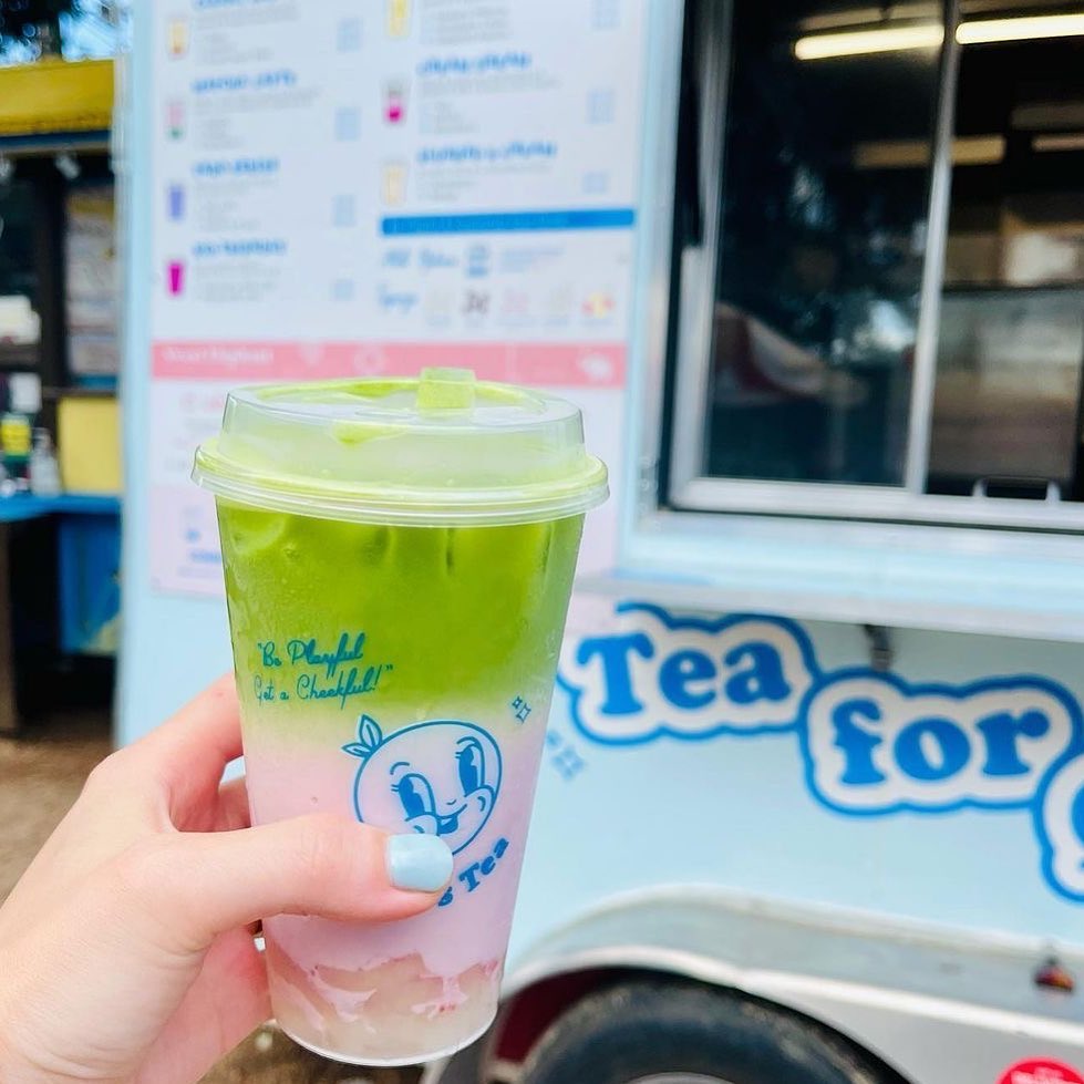 A hand holding up a clear plastic glass of a green and pink liquid in front of a pastel food truck with a partial sign reading “Tea for”.