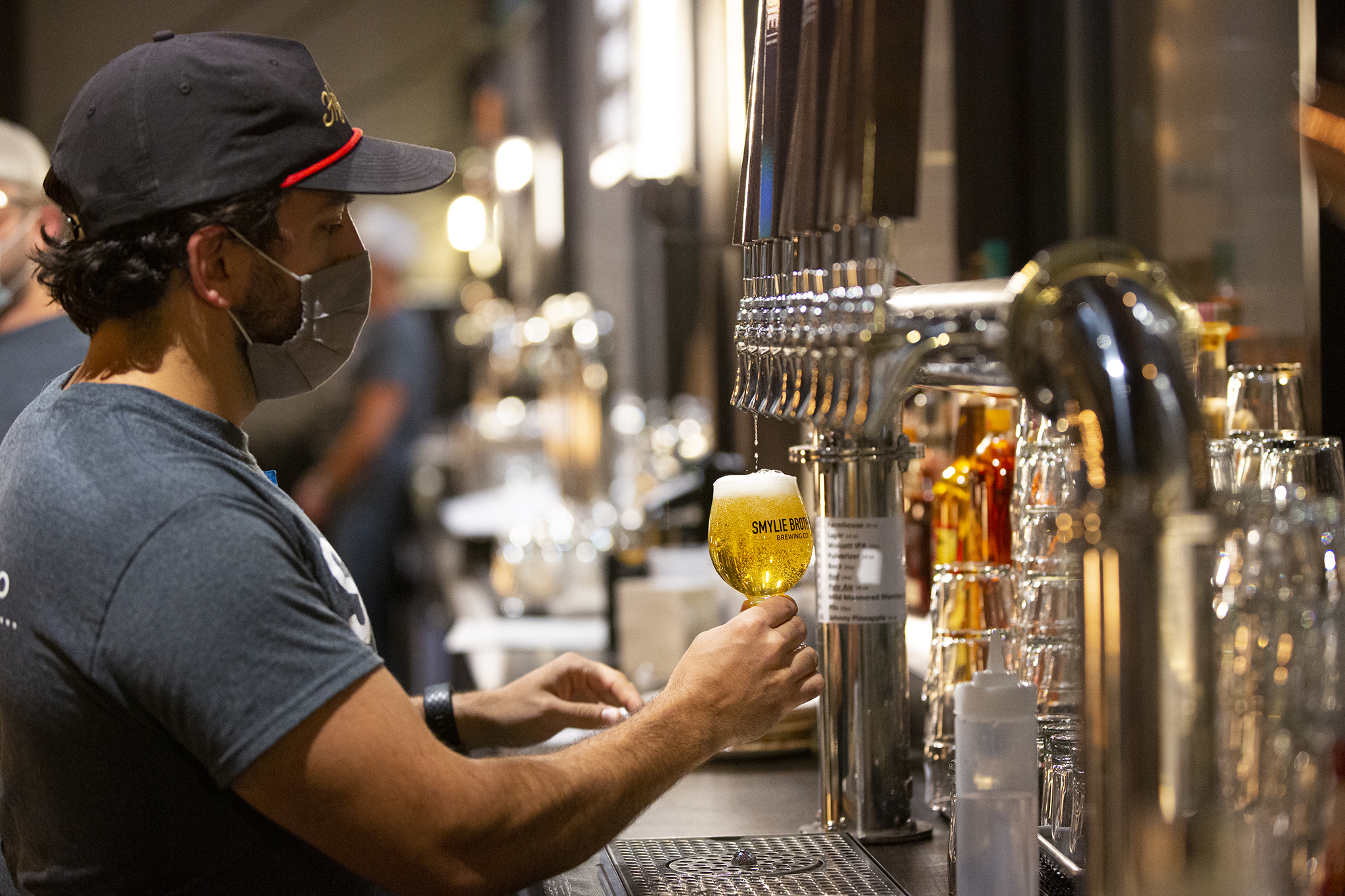 A bartender in a baseball cap and face mask pulls a beer from the tap