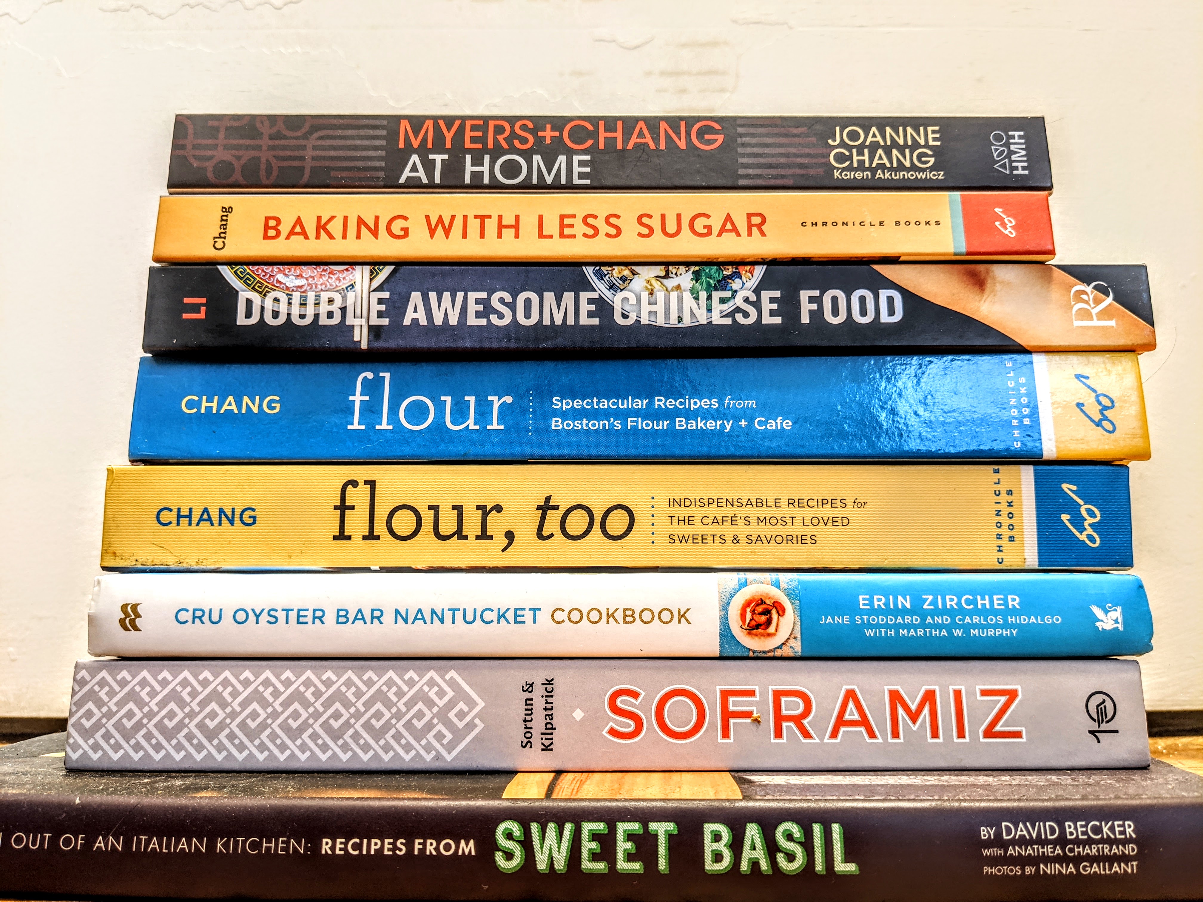 A stack of eight cookbooks, spines facing the camera, with titles including Sweet Basil, Soframiz, Double Awesome Chinese Food, and more.