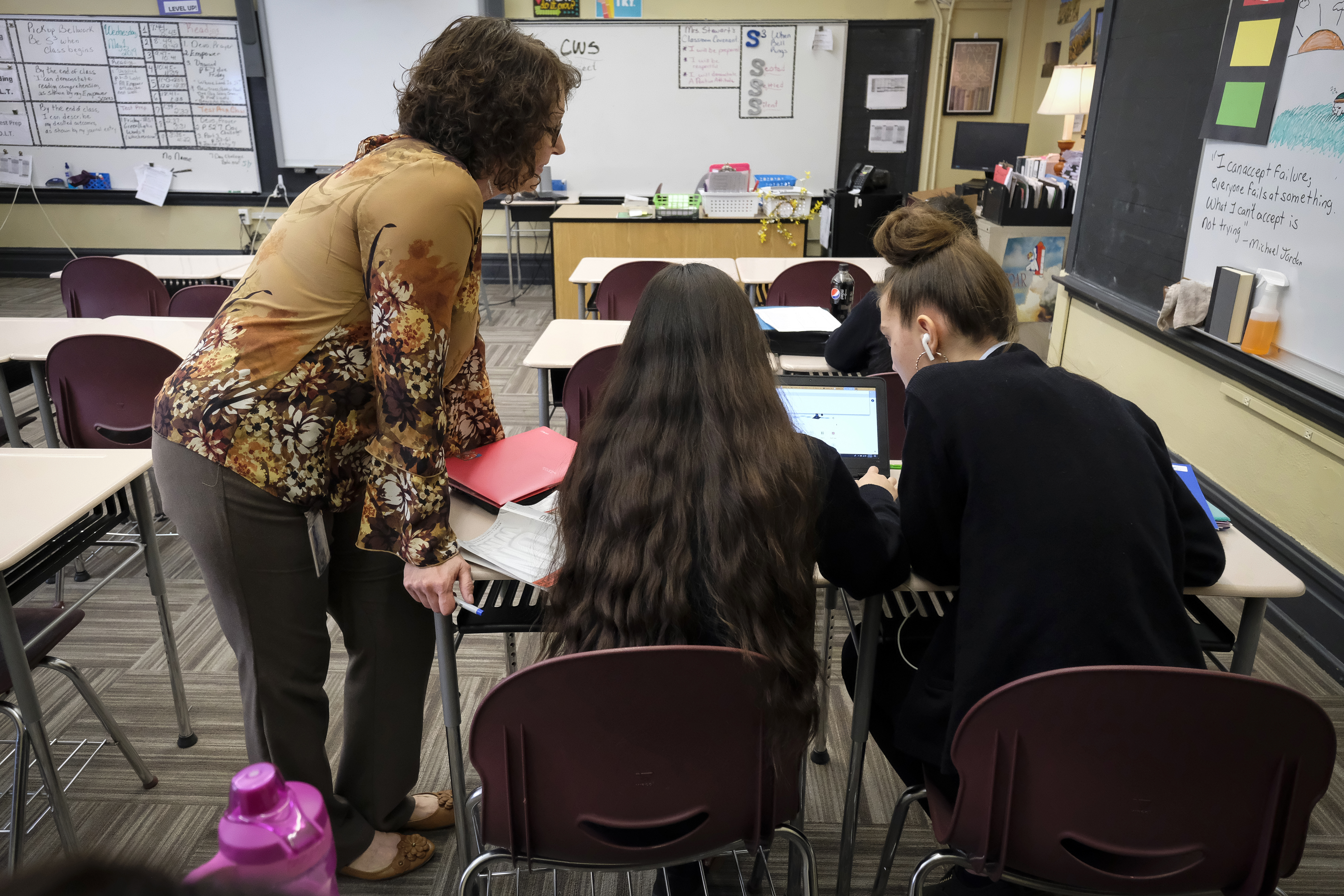 In a classroom, two students work together on a laptop while a teacher looks on.