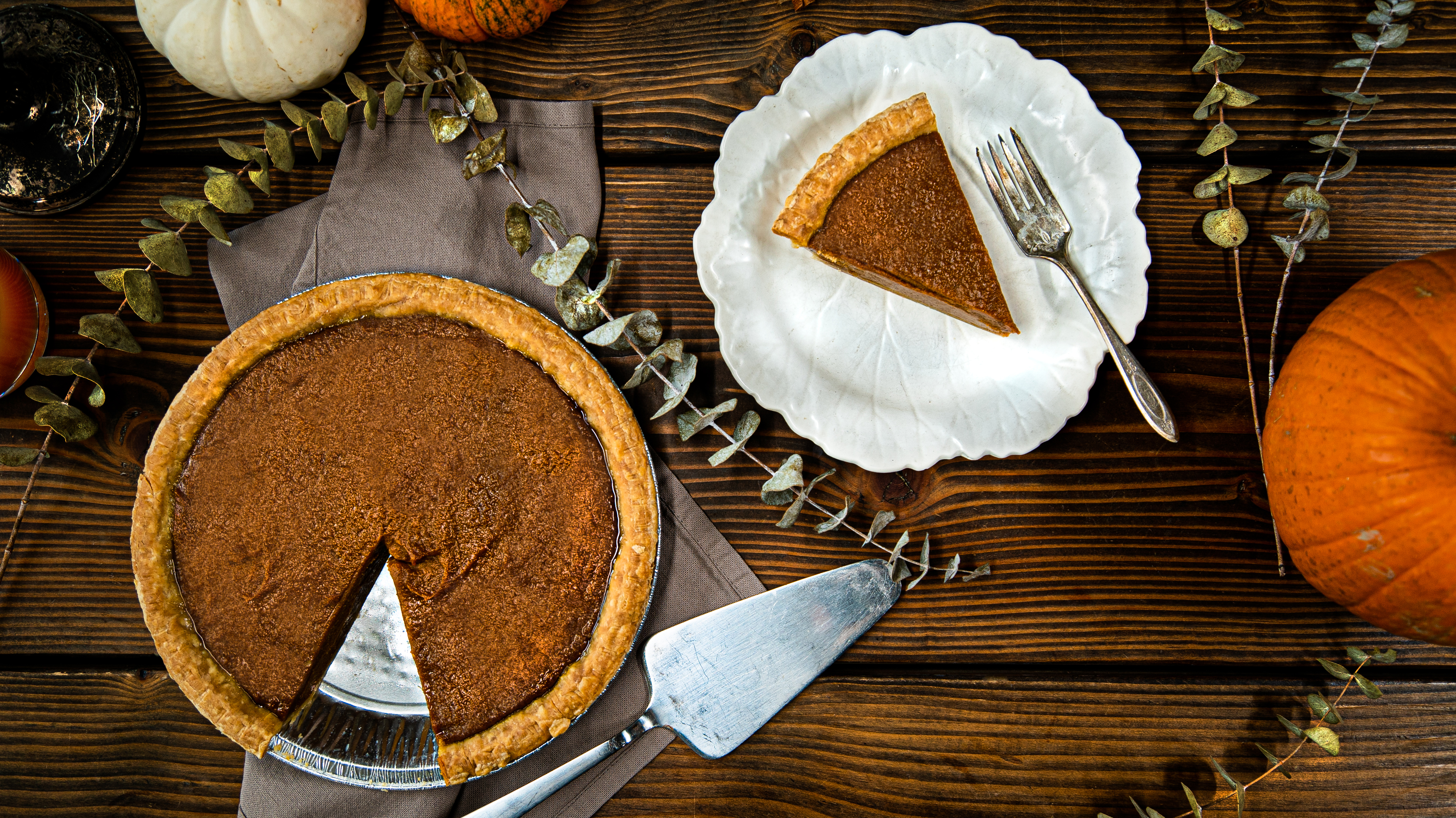 An overhead shot of a sweet potato pie with one slice taken out on a plate with pumpkins and eucalyptus scattered around.