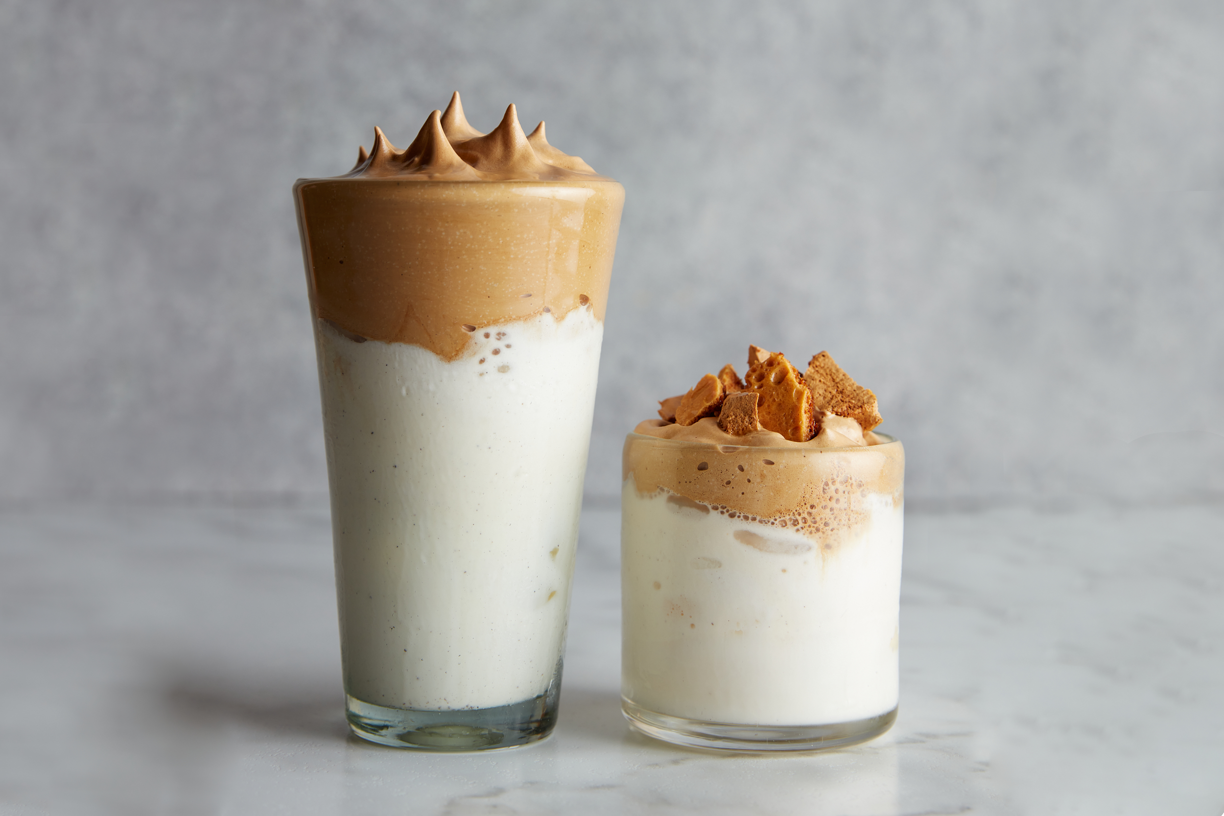 One tall glass and one short glass of dalgona milkshake sit next to each other on a marble surface.