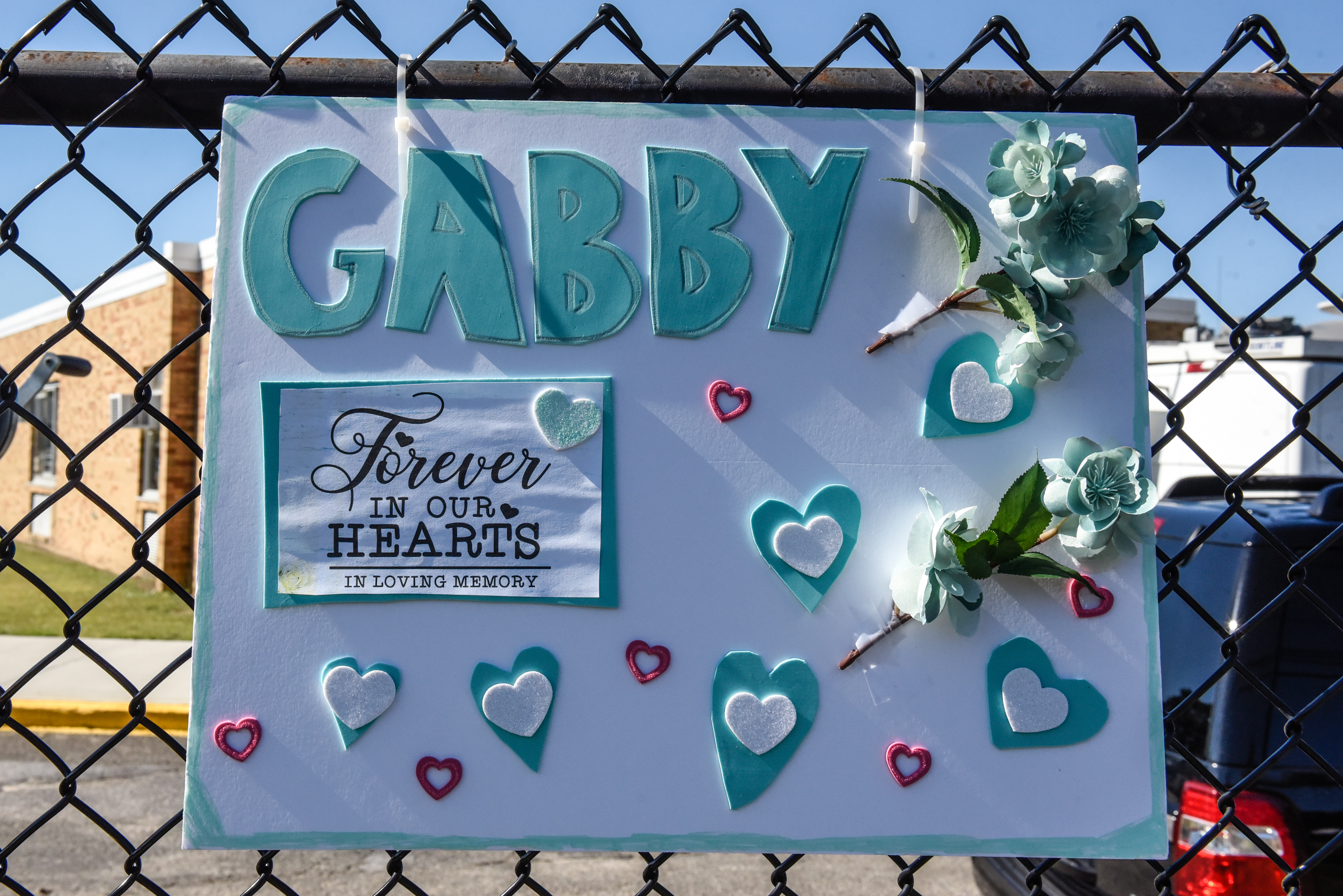 A homemade sign covered in hearts and flowers reads, “Gabby, forever in our hearts.”