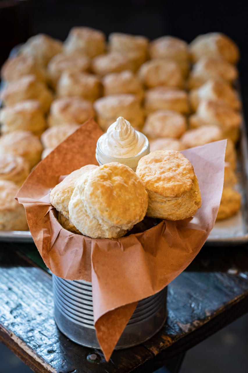 Several plump buttermilk biscuits are stacked inside a big tin can lined with brown paper. There’s a small side of whipped butter.