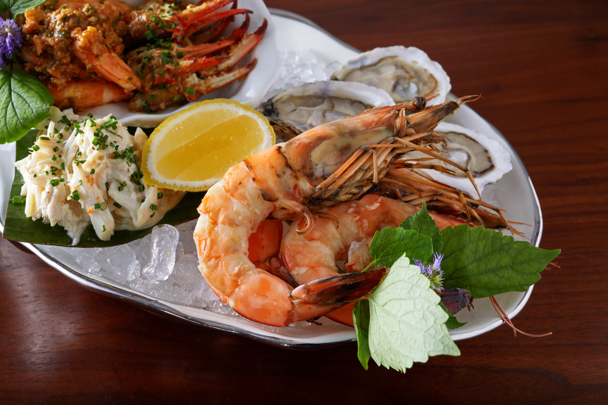 A silver platter topped with crushed ice holding a mix of large steamed prawns, crab claws, oysters on the half shell, and smoked fish dip.