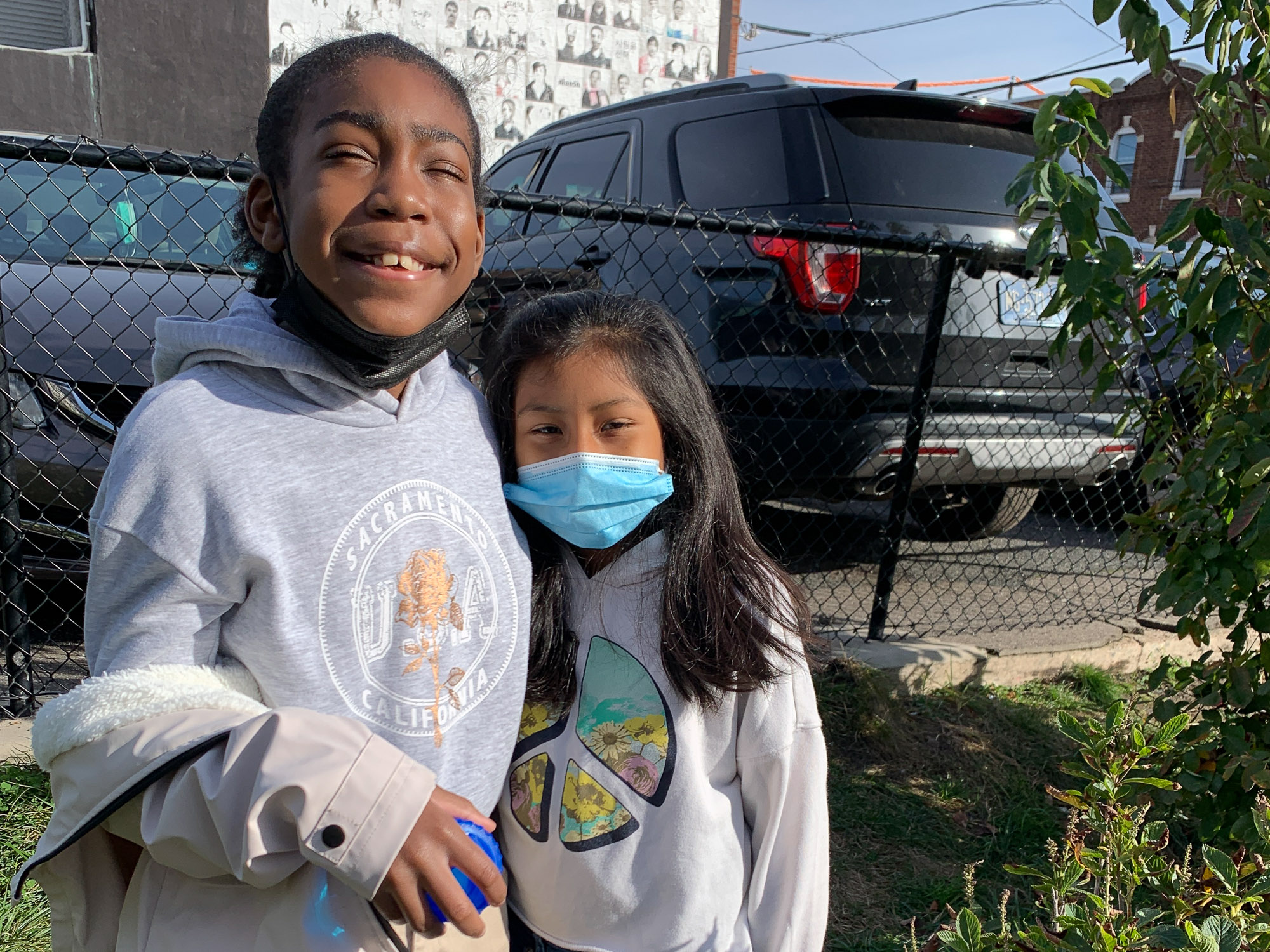 Two fourth grade students wearing hoodies pose together for a portrait in a small green space next to a parking lot.